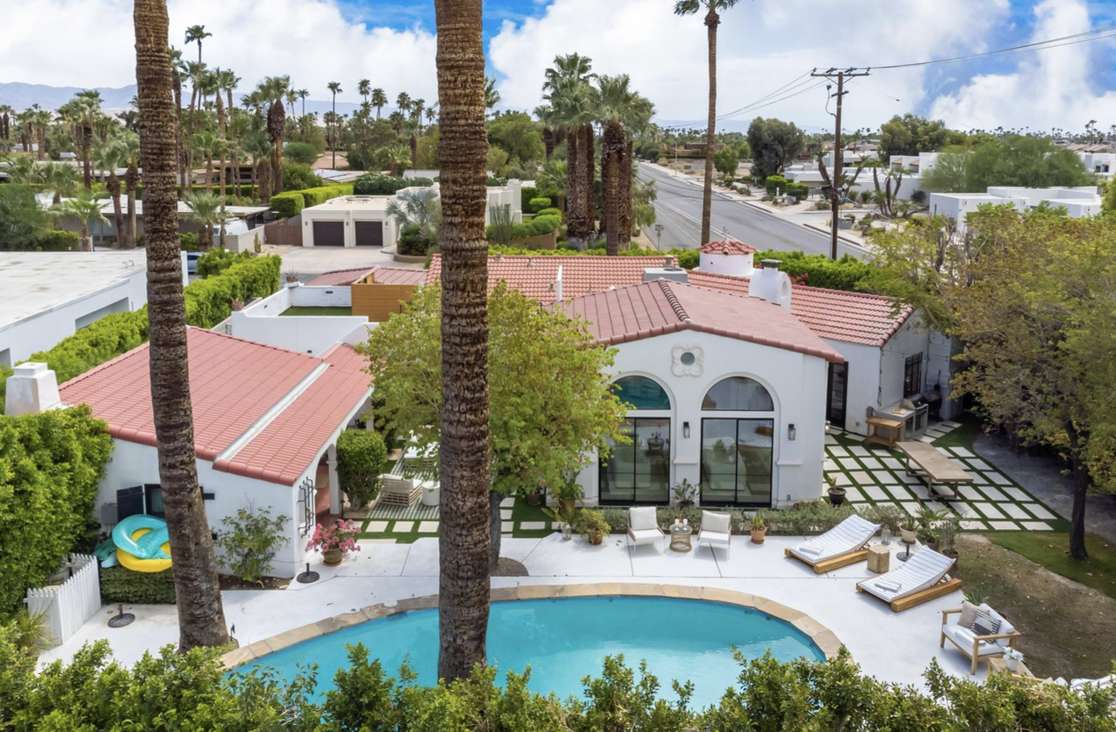 A single-story house with a red tile roof is surrounded by palm trees and features a pool with lounge chairs in the backyard.