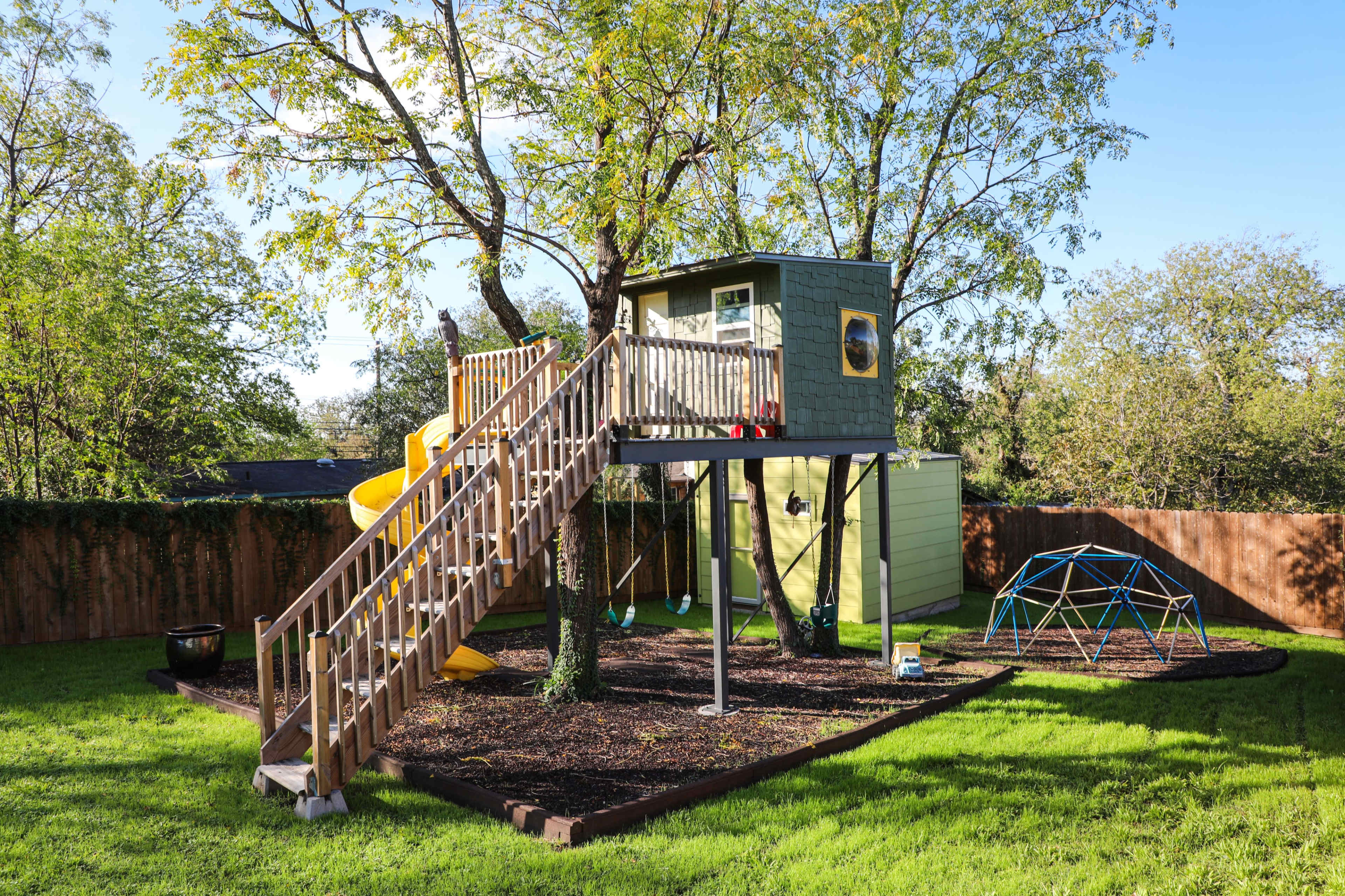 The image shows a backyard with a treehouse on stilts, featuring a slide and a wooden ladder, surrounded by a grassy lawn and a play structure in the background.
