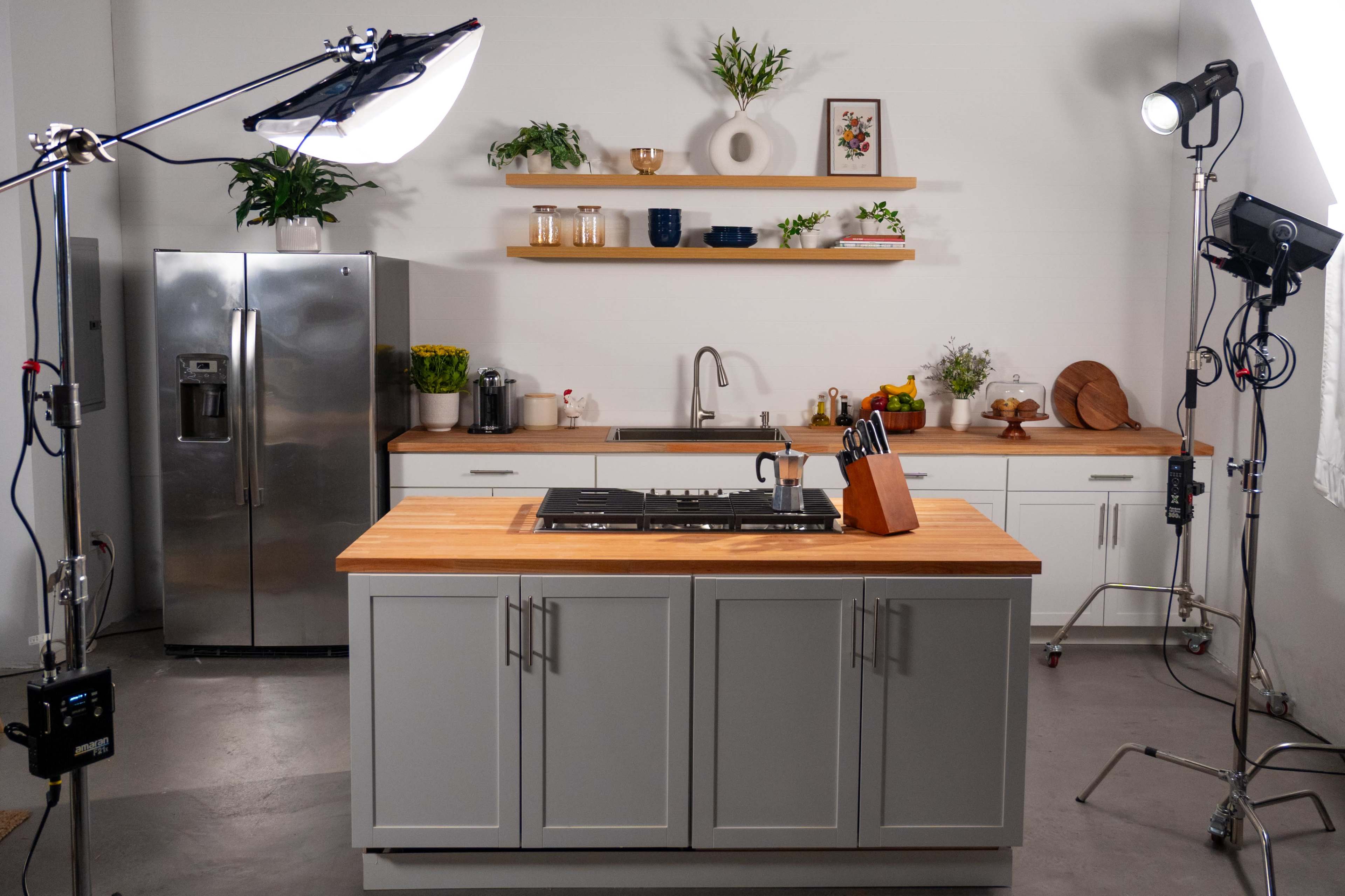 A modern kitchen setup features a central island with a sink, a stainless steel refrigerator, and shelves displaying various kitchen items.