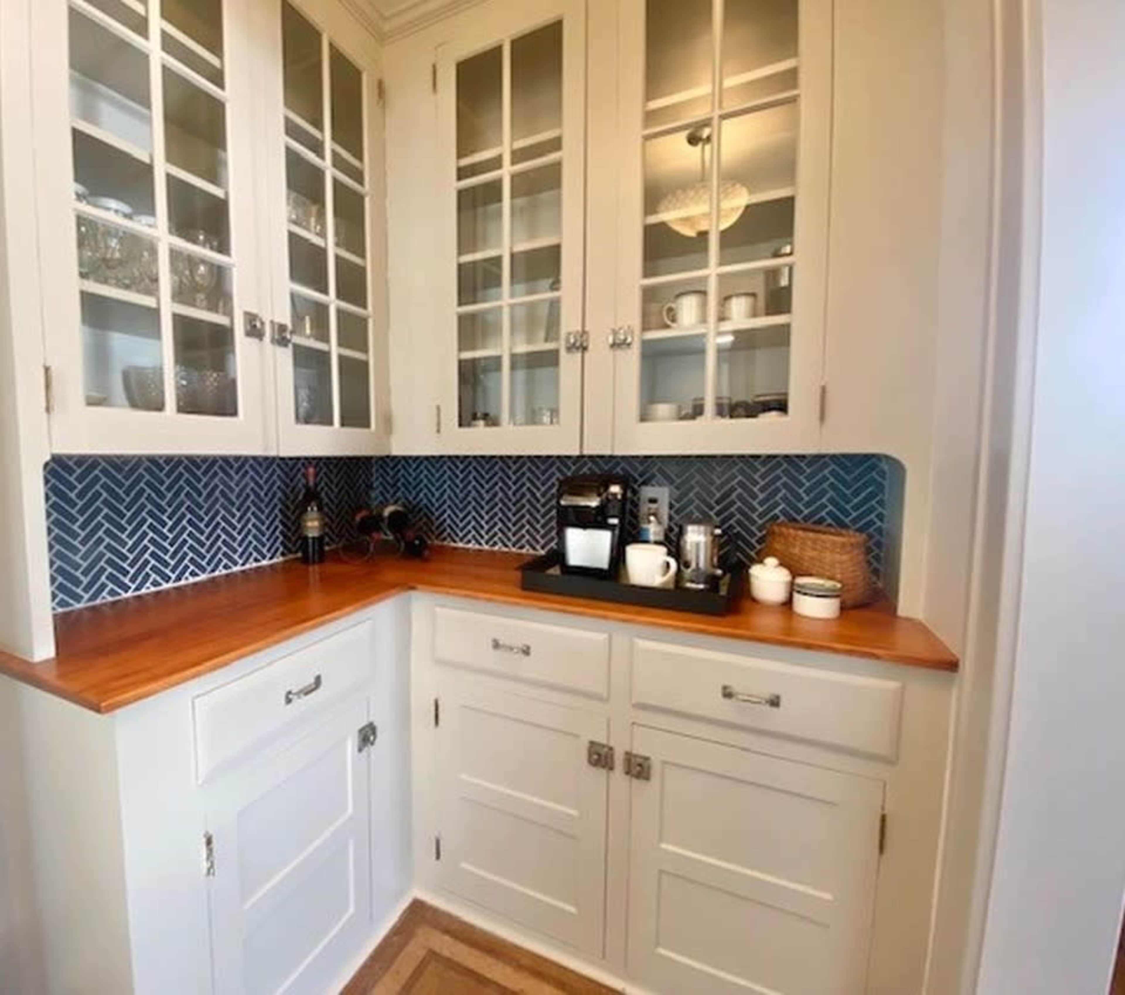 The image shows a kitchen nook featuring white cabinets with glass doors, a wooden countertop, and a coffee station against a blue herringbone backsplash.