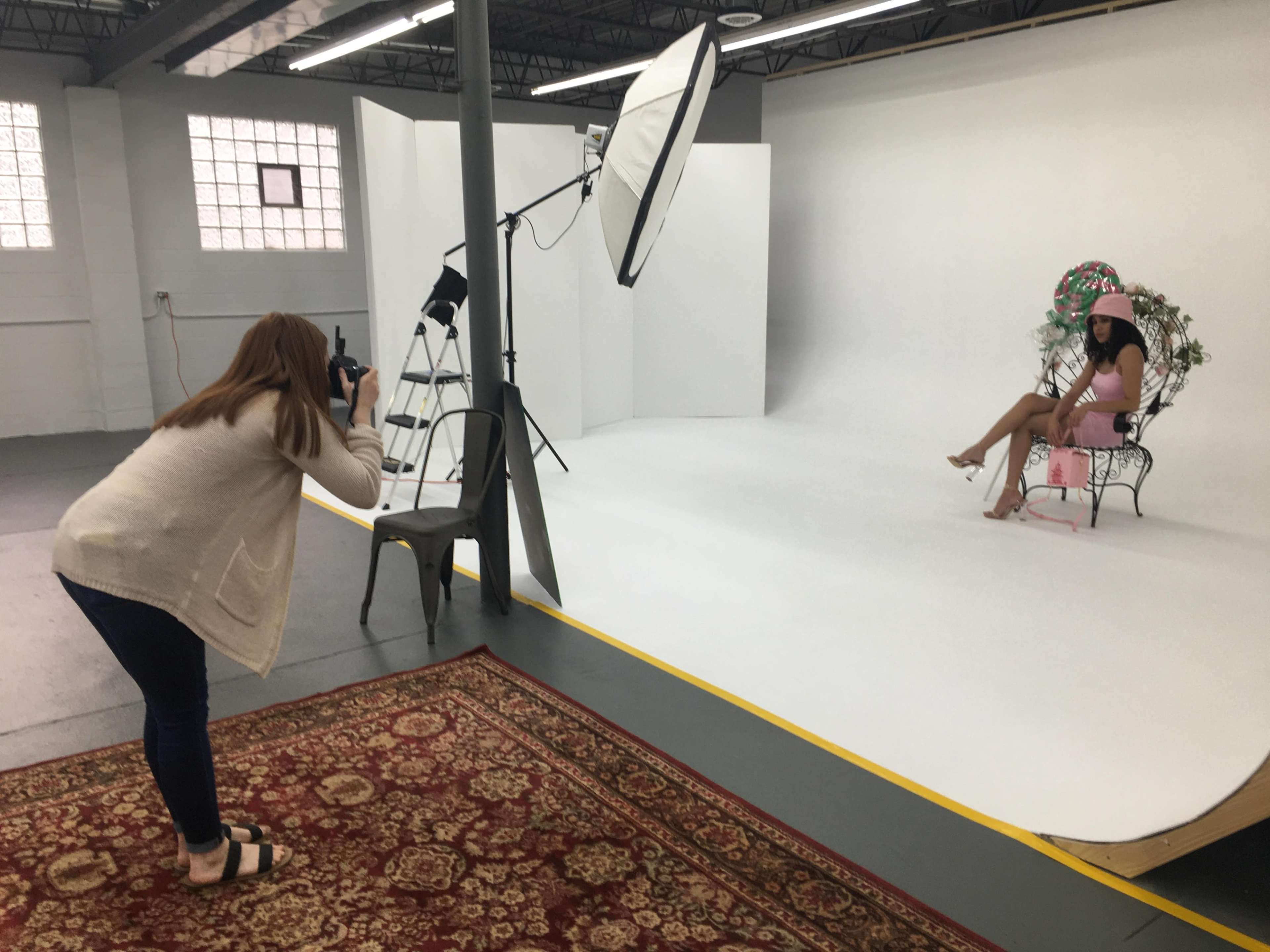A photographer captures a model seated in a chair against a white backdrop in a studio setting.