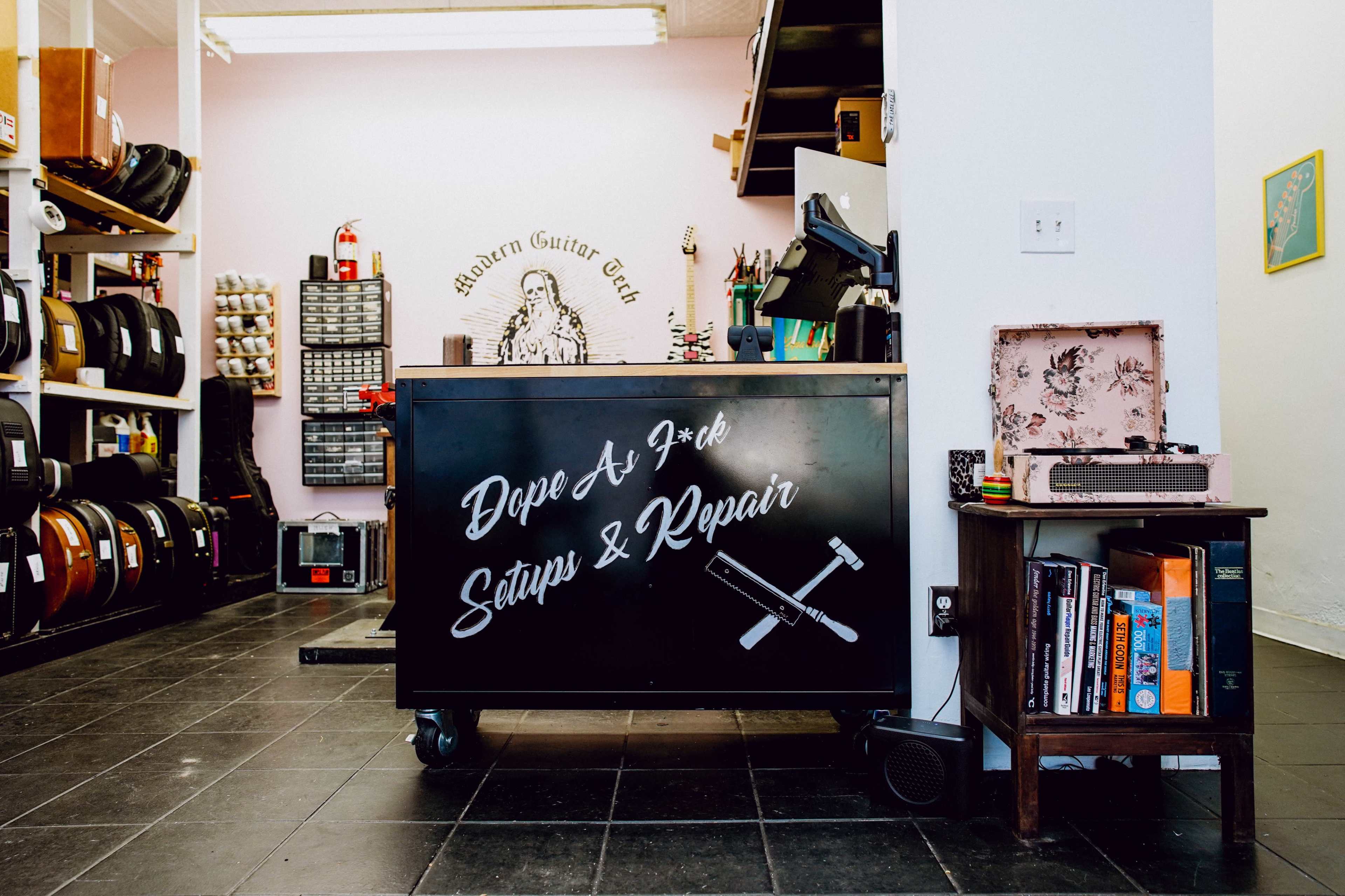The image shows a workspace with a black cart displaying "Dope As F**k Setups & Repair" and various musical instruments and equipment in the background.