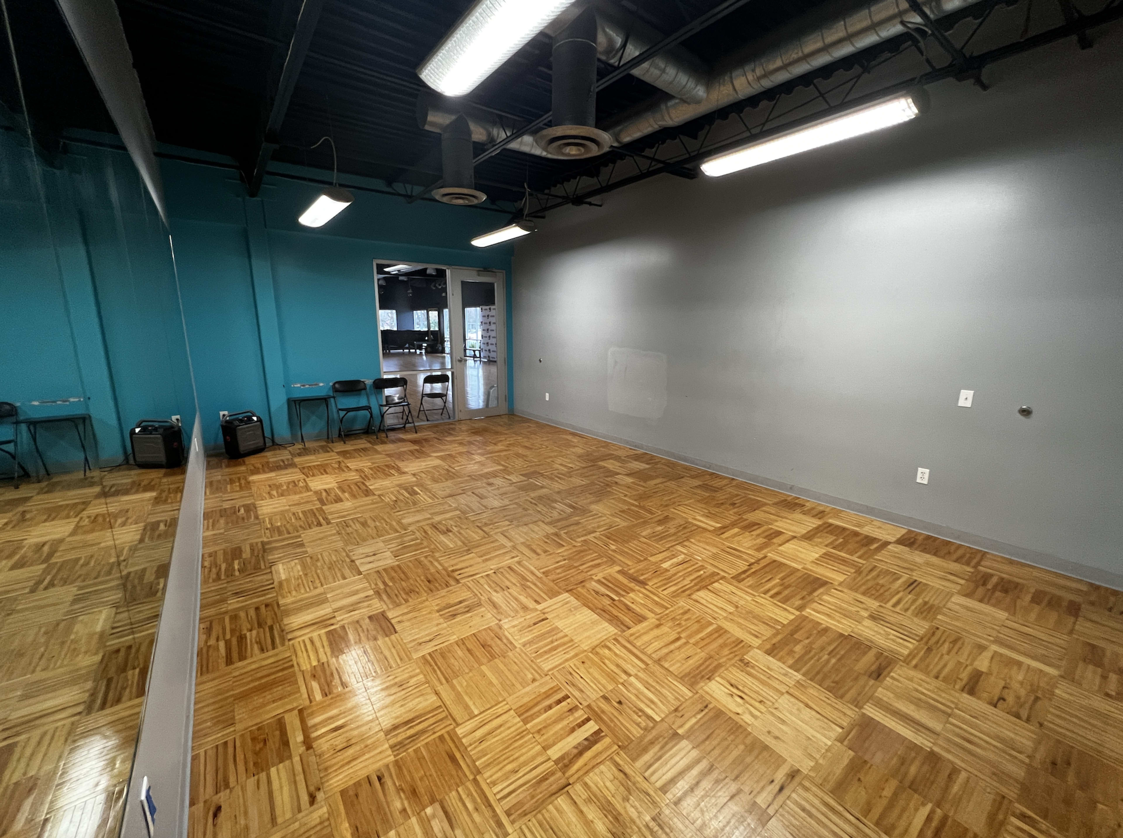 The image shows an empty room with wooden flooring, a mirrored wall on one side, and teal-colored and gray walls.