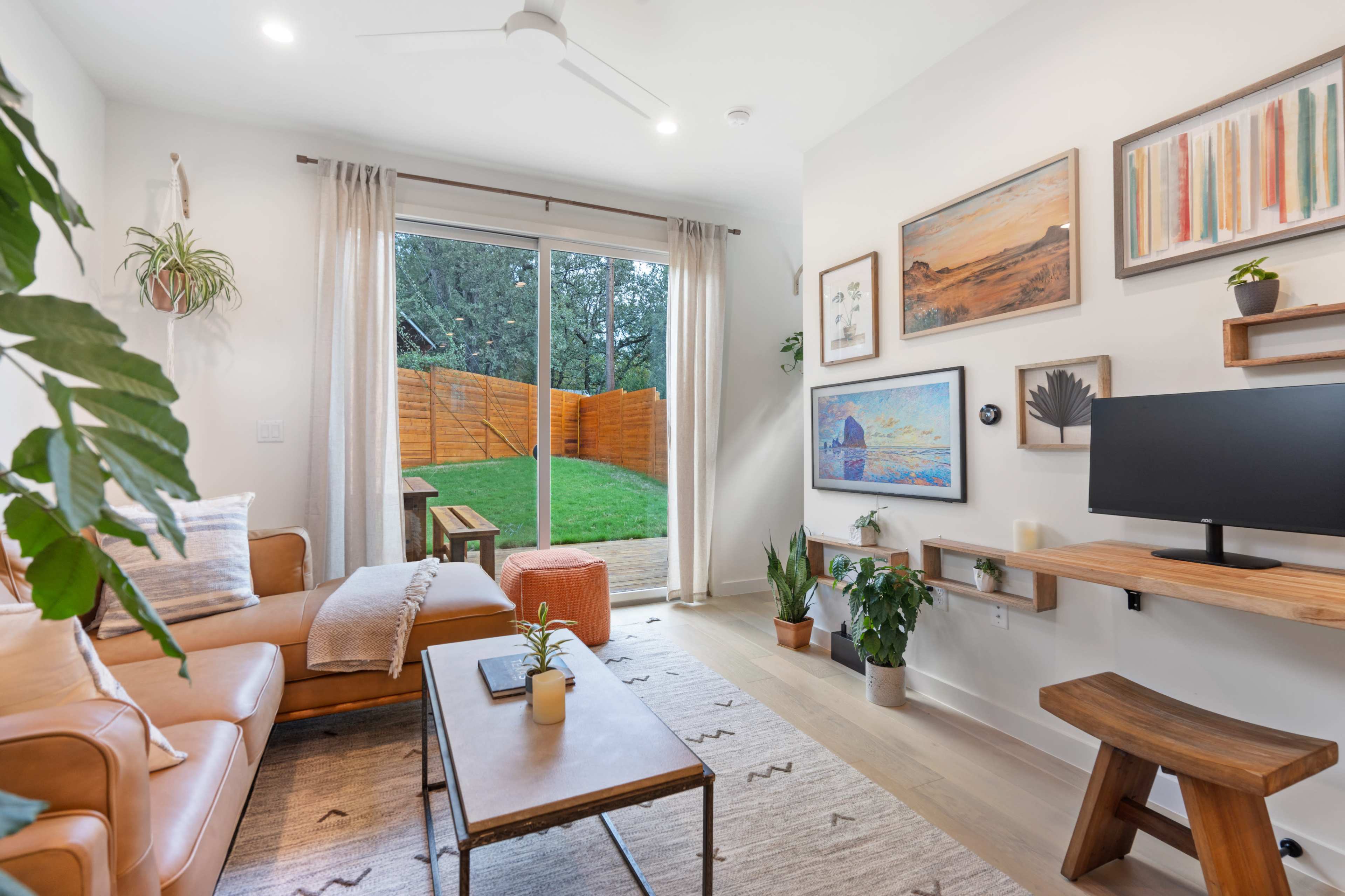 The image shows a modern living room with a leather sofa, a wooden coffee table, and a workspace mounted on the wall, overlooking a garden through sliding glass doors.