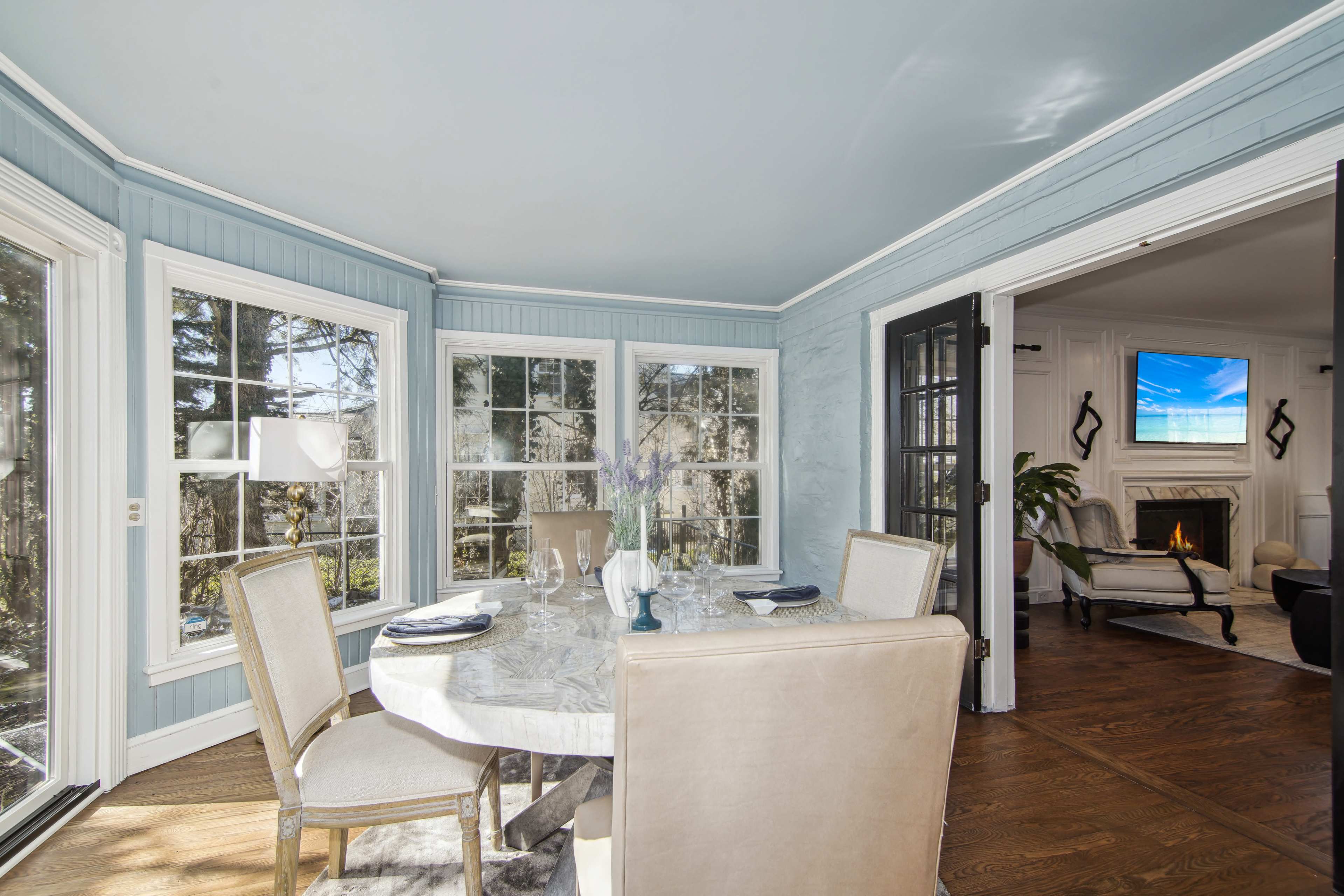 A dining area with a round table set for a meal, surrounded by large windows and opening to a living room with a fireplace.