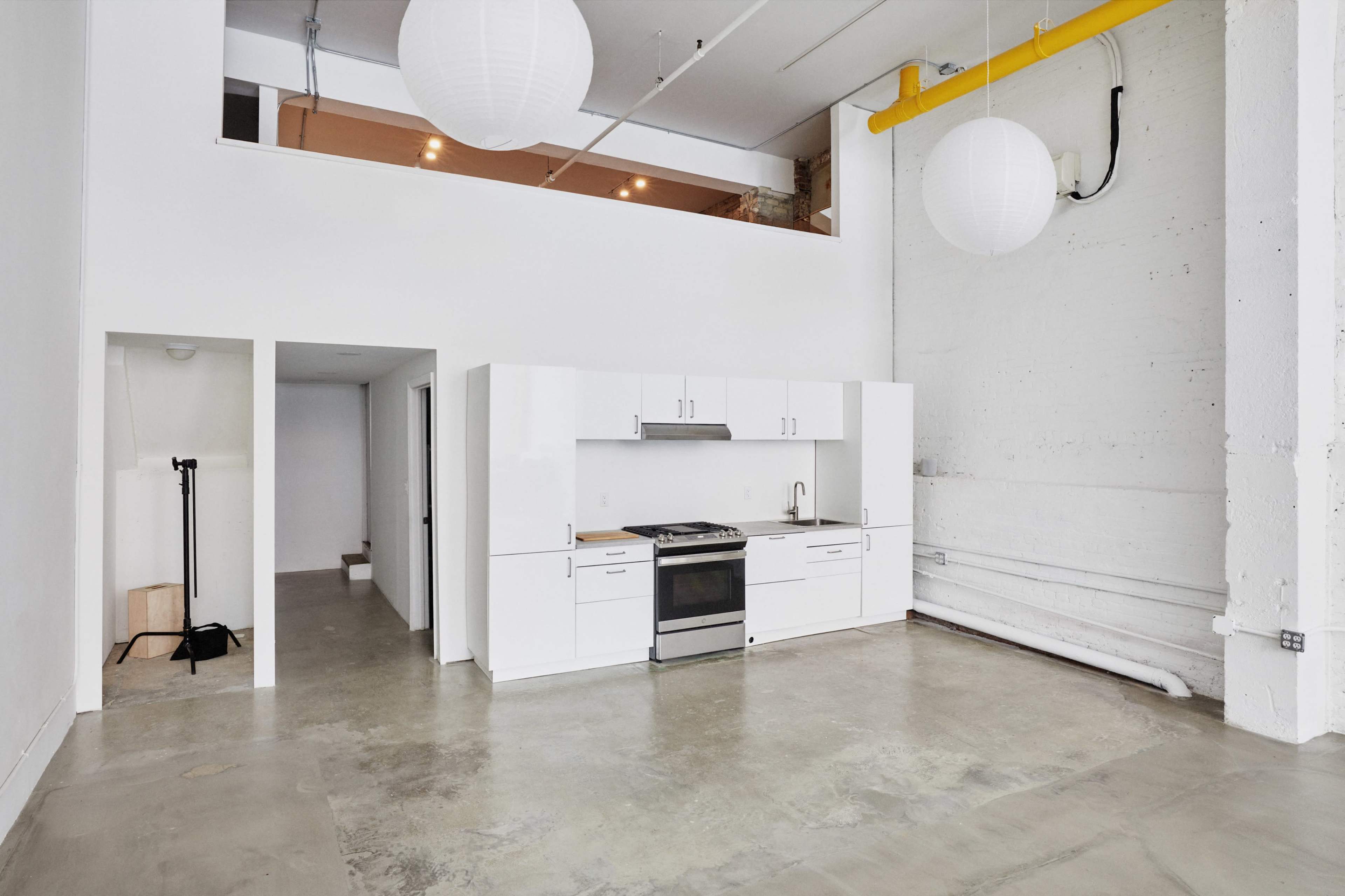 A modern, open-concept kitchen area with white cabinets and a concrete floor, alongside a large, bright space featuring pendant lights.
