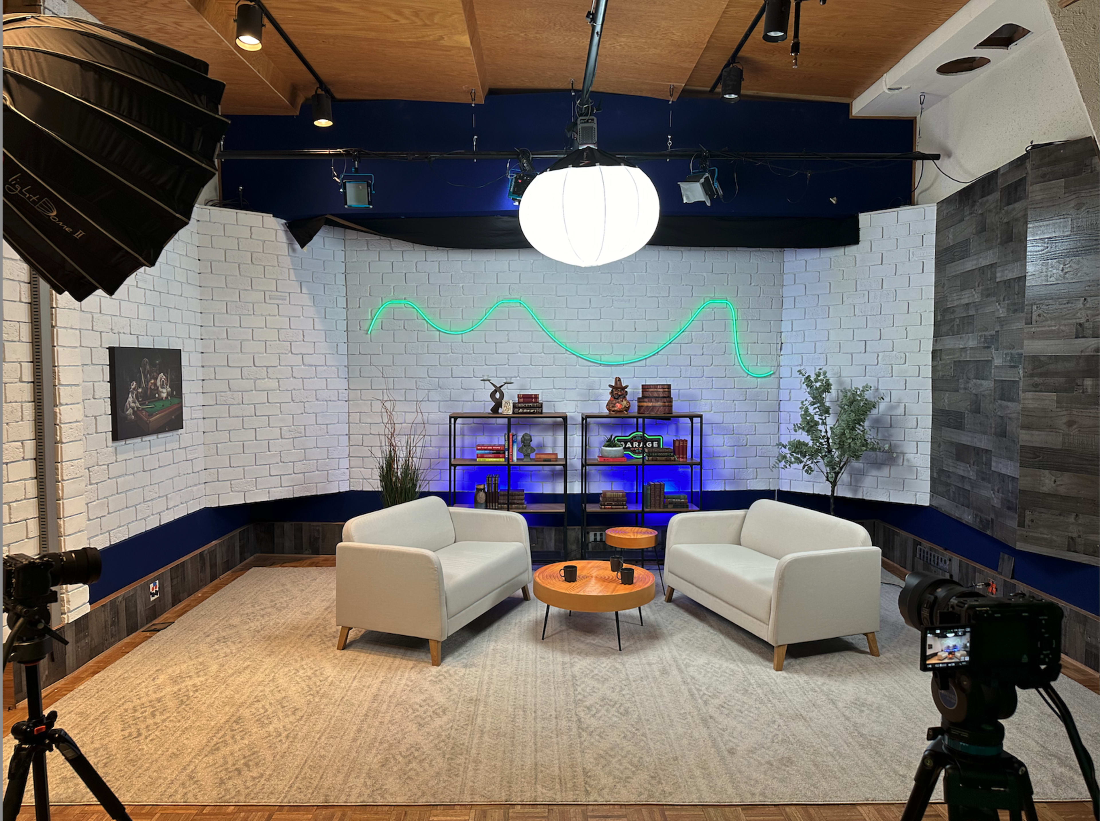 The image shows a modern studio setup featuring two white sofas facing each other, a round wooden coffee table in between, and a decorative background with shelves and ambient lighting.