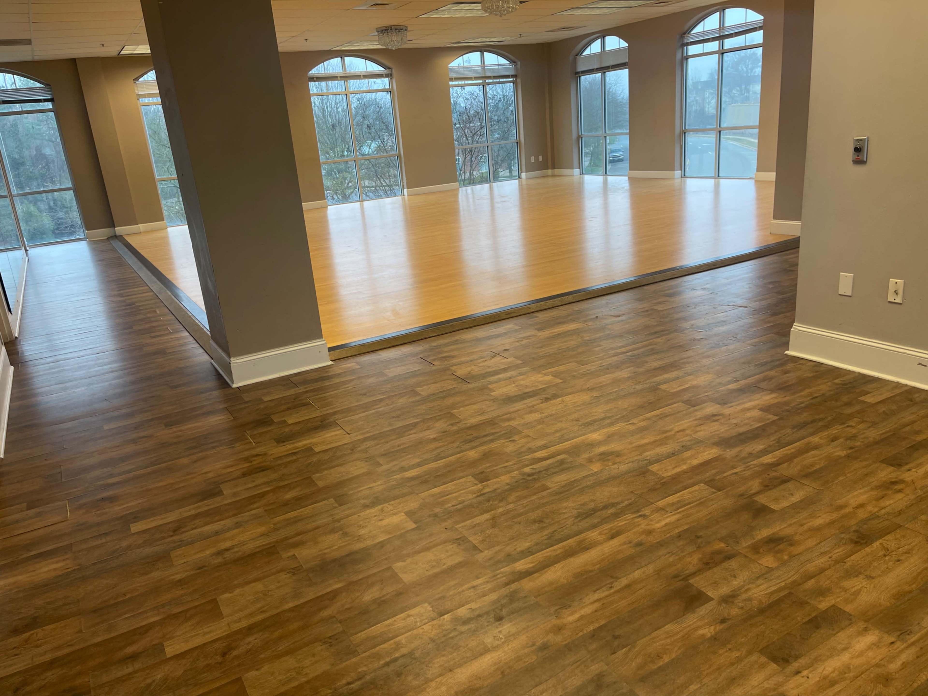 A spacious room with large windows, featuring a wooden floor and a smooth, light-colored dance area in the center.