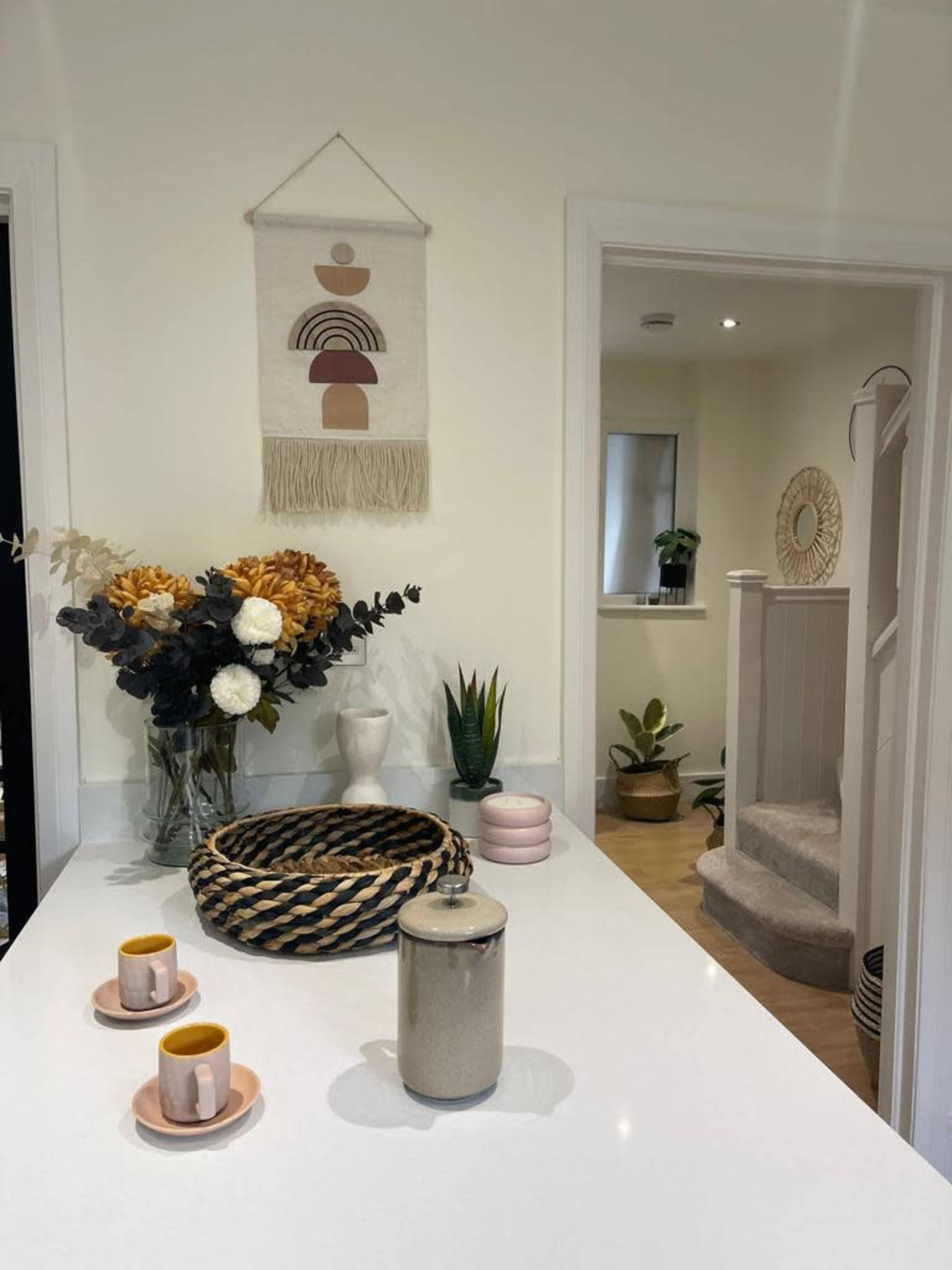 The image shows a modern kitchen counter with a decorative arrangement of flowers, a woven basket, decorative jars, and an opening leading to a staircase and another room.