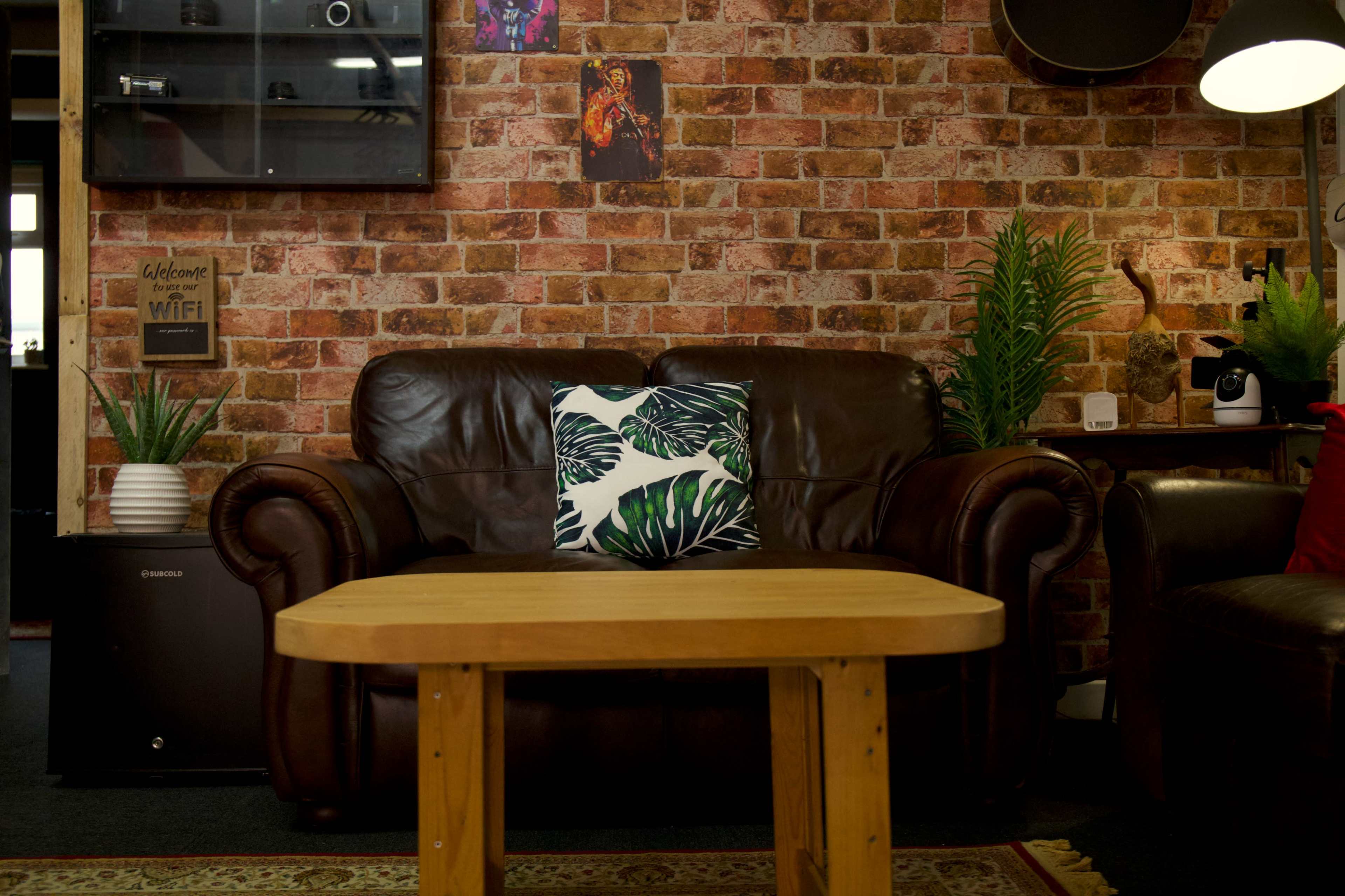 A brown leather sofa with a leafy decorative pillow is positioned in front of a wooden coffee table against a brick-patterned wall adorned with plants and wall art.
