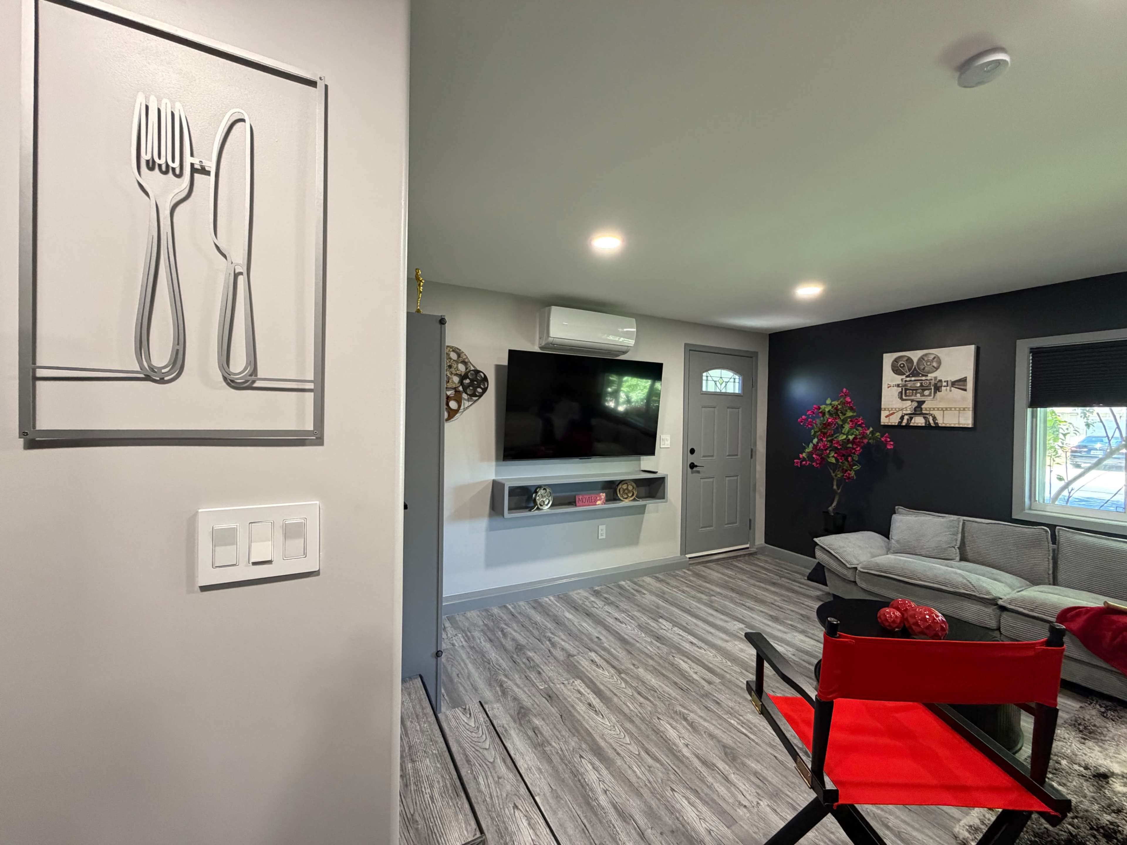 The image shows a modern living room with a grey couch, a wall-mounted TV, and a decorative wall featuring a fork and knife artwork.