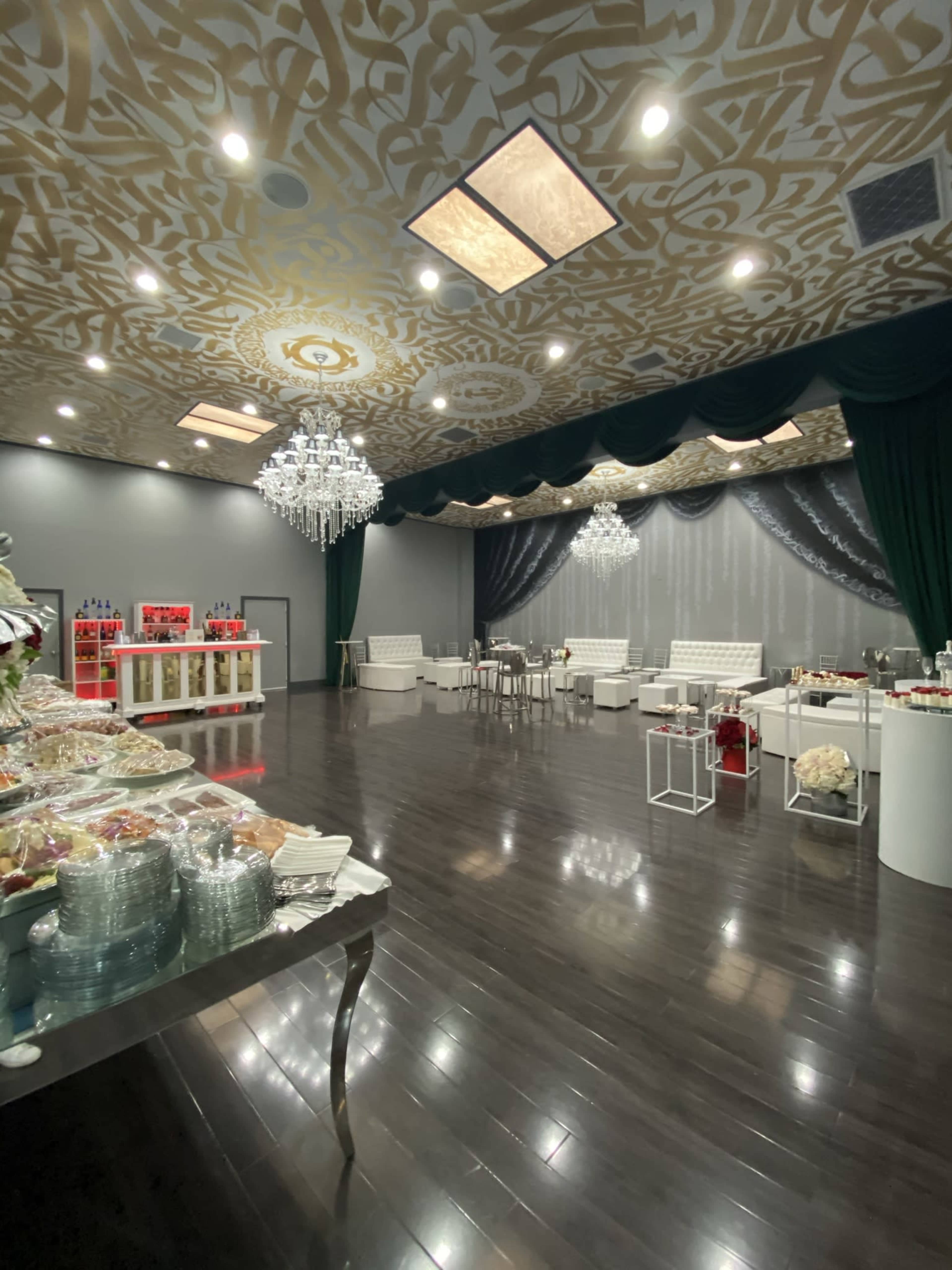 A spacious event hall with elegant decor, a buffet table on the left, and seating arrangements featuring white furniture and decorative chandeliers.