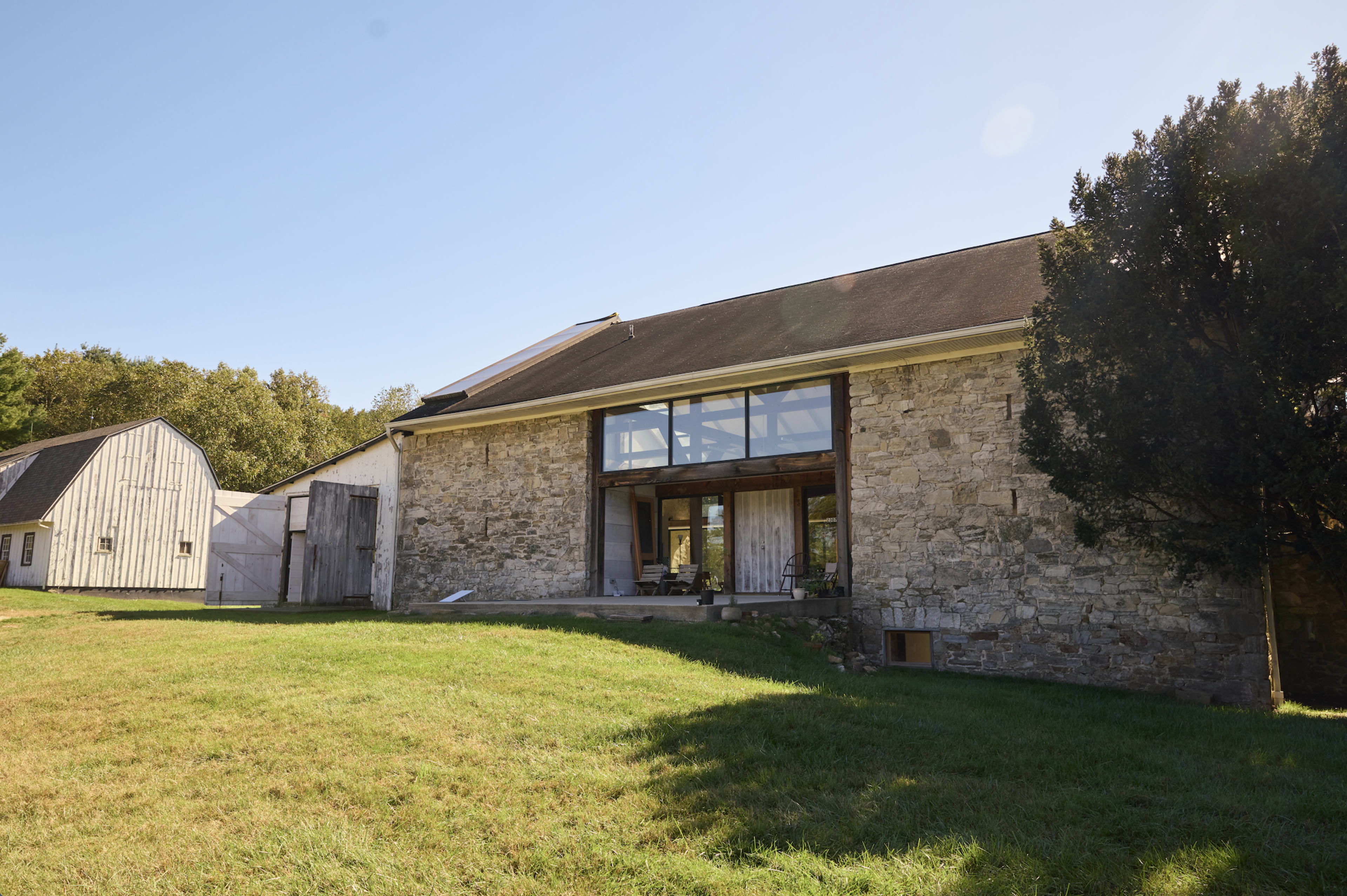 A stone building with a large glass window is situated on a grassy area, surrounded by trees and a nearby wooden structure.