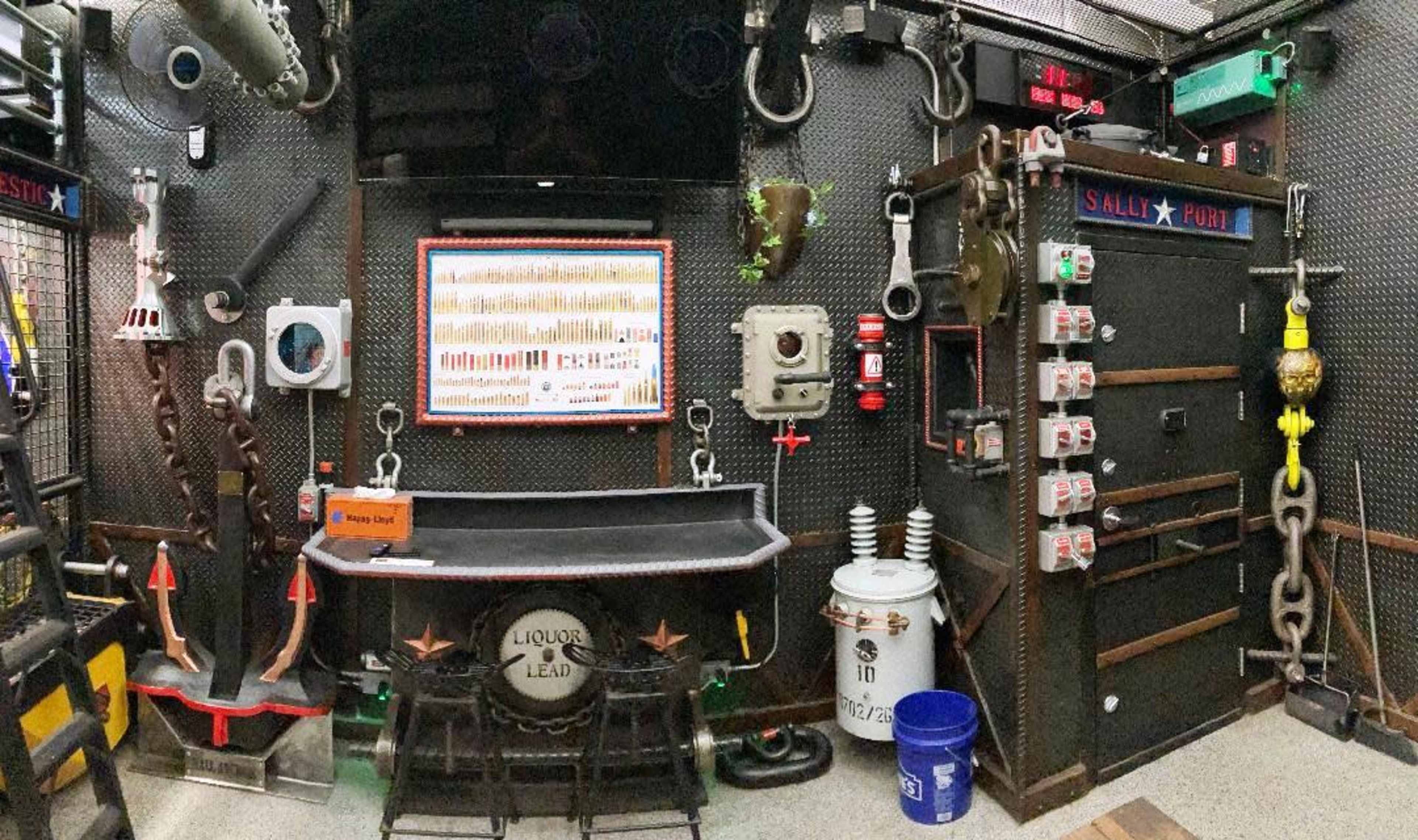 The image shows a wall filled with industrial tools and equipment, including chains, a large door, and various metallic objects, arranged in a dimly lit space.