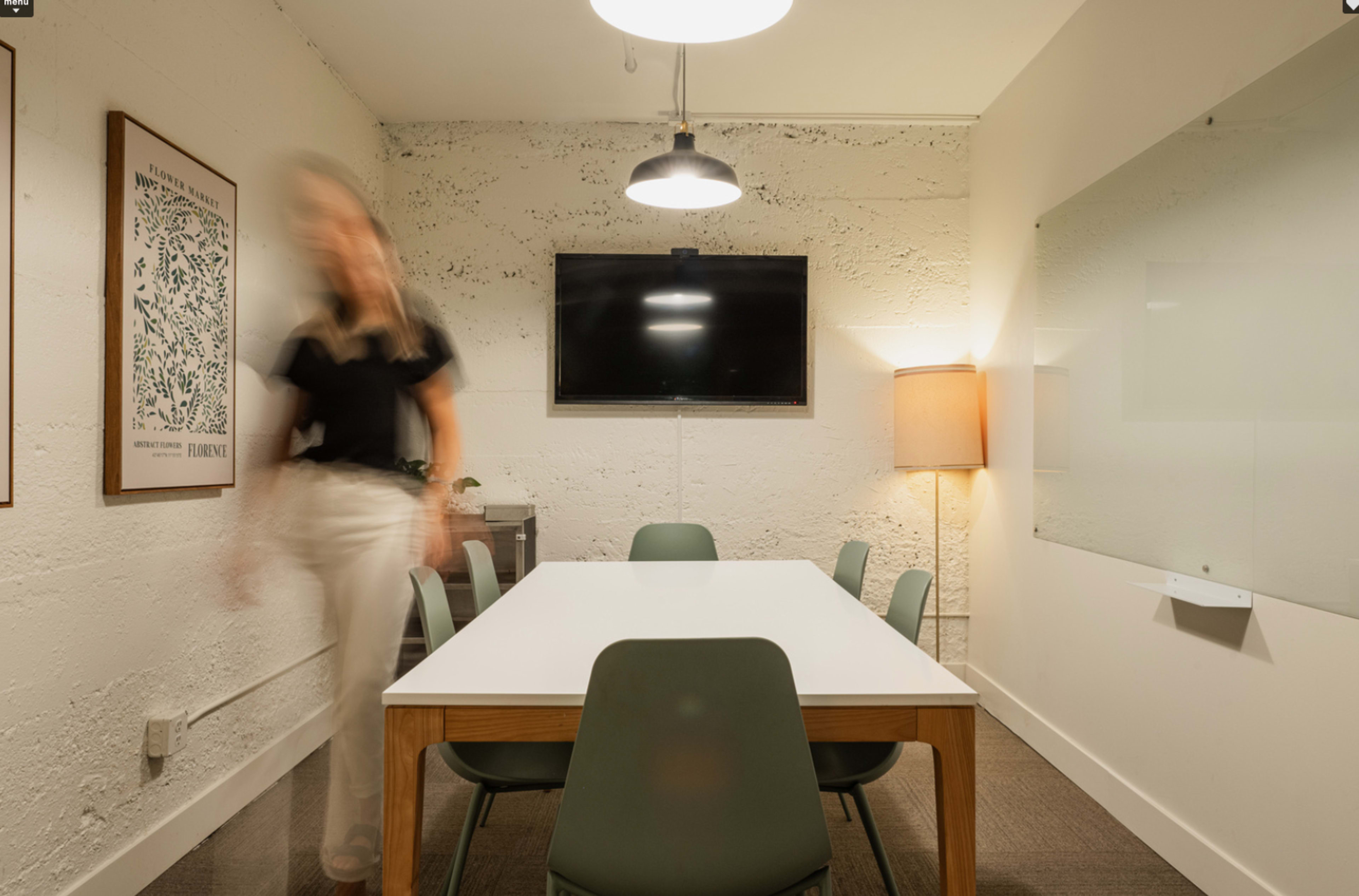 A modern meeting room features a white table surrounded by green chairs, a television on the wall, and a lamp in the corner.