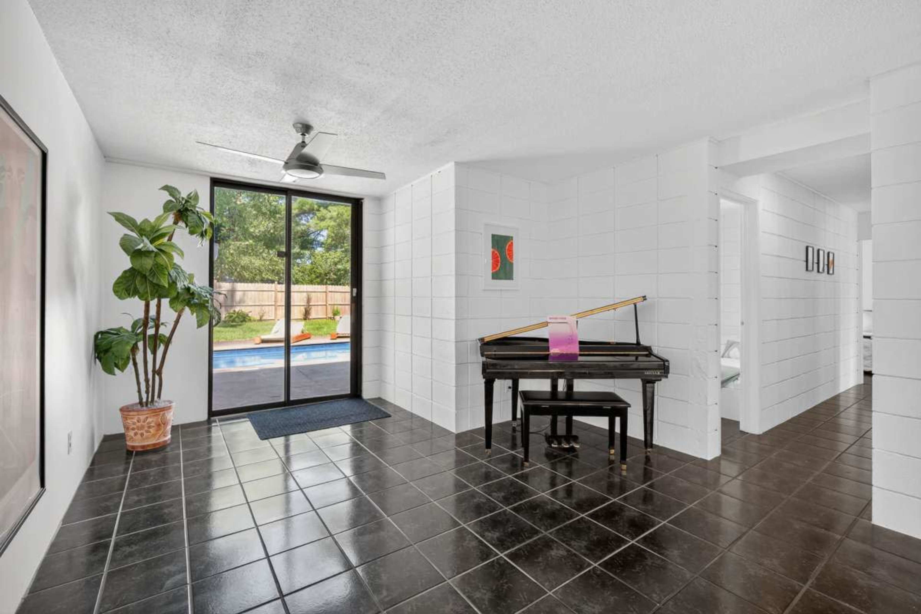 The room features a black tile floor, a grand piano, a large window with a view of a pool, and a potted plant in the corner.