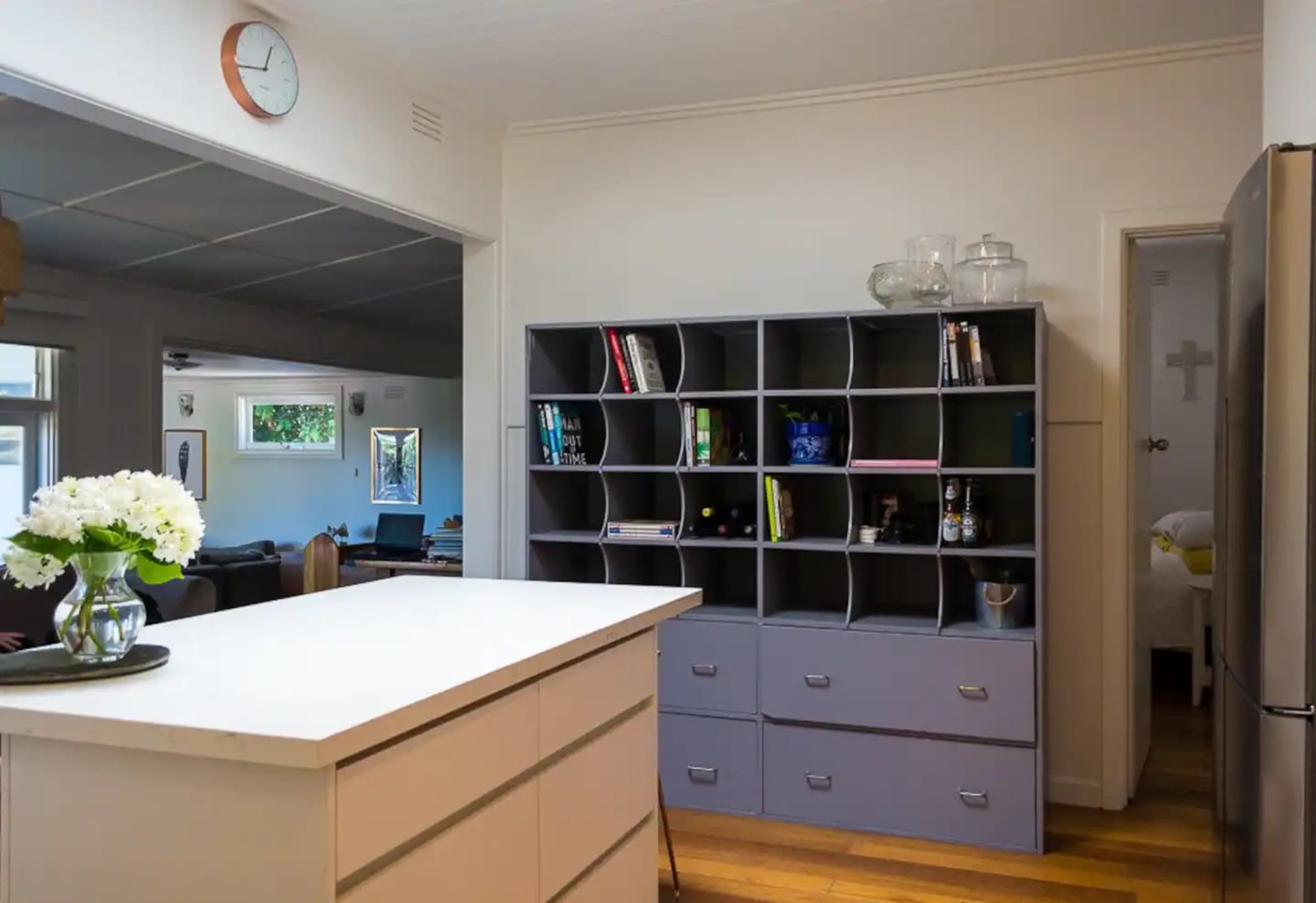 A modern kitchen area featuring a white island with a dark gray bookcase filled with books and decorative items, alongside a stainless steel refrigerator.