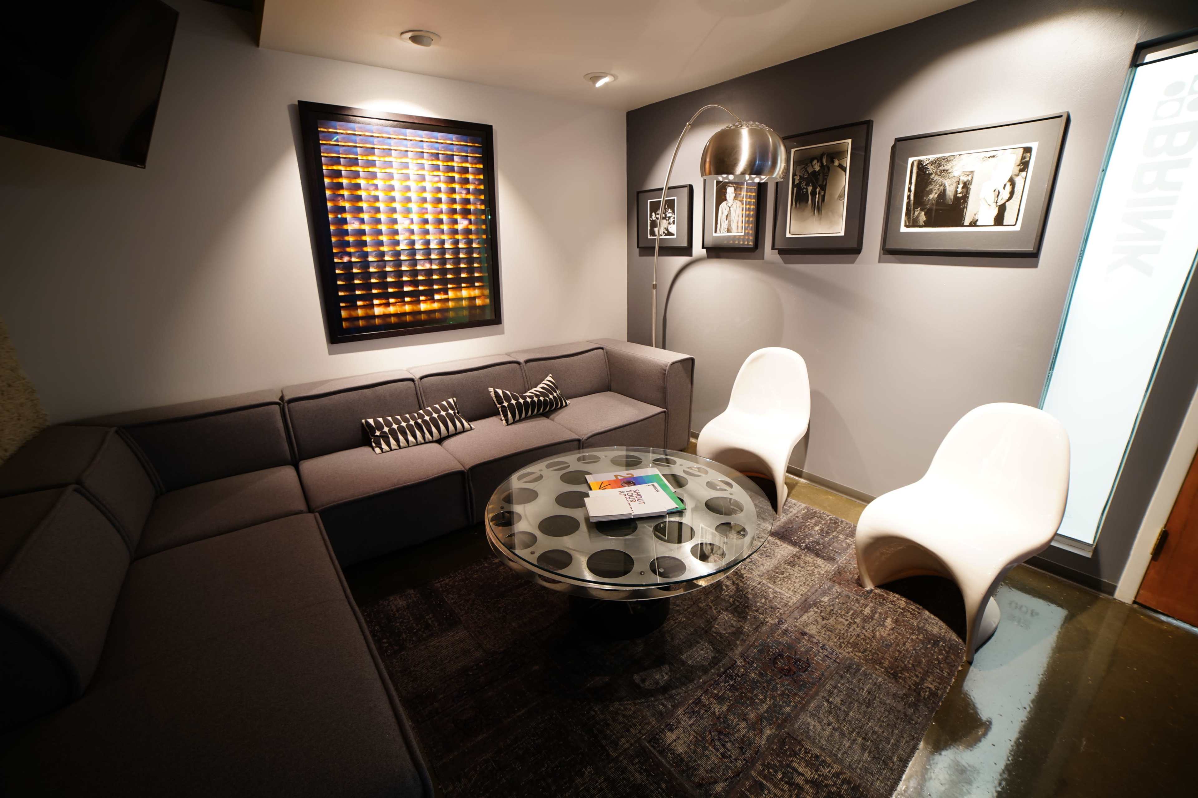 A modern lounge area with a sectional sofa, a round glass coffee table, and two white chairs, accented by framed black-and-white photographs on the walls.