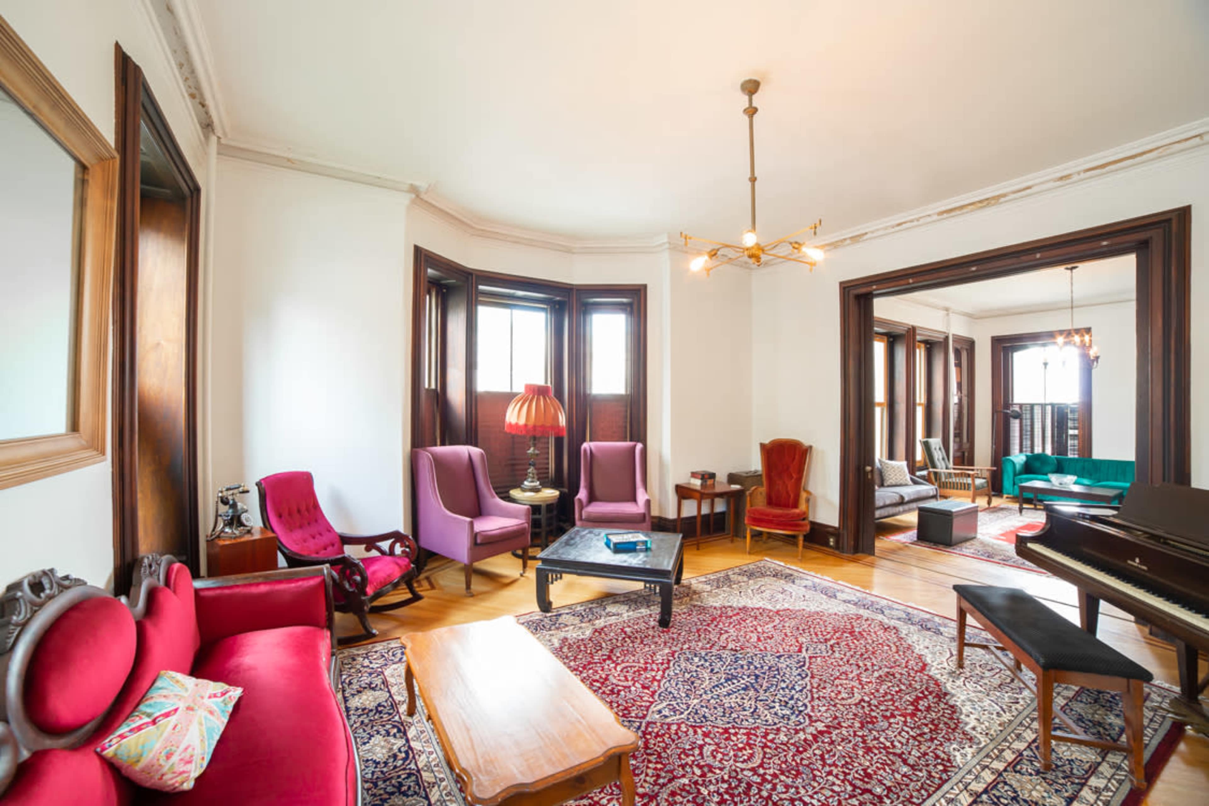 A well-furnished living room with a mix of seating areas, including purple and red chairs, a wooden coffee table, and a piano, all arranged around a patterned rug.