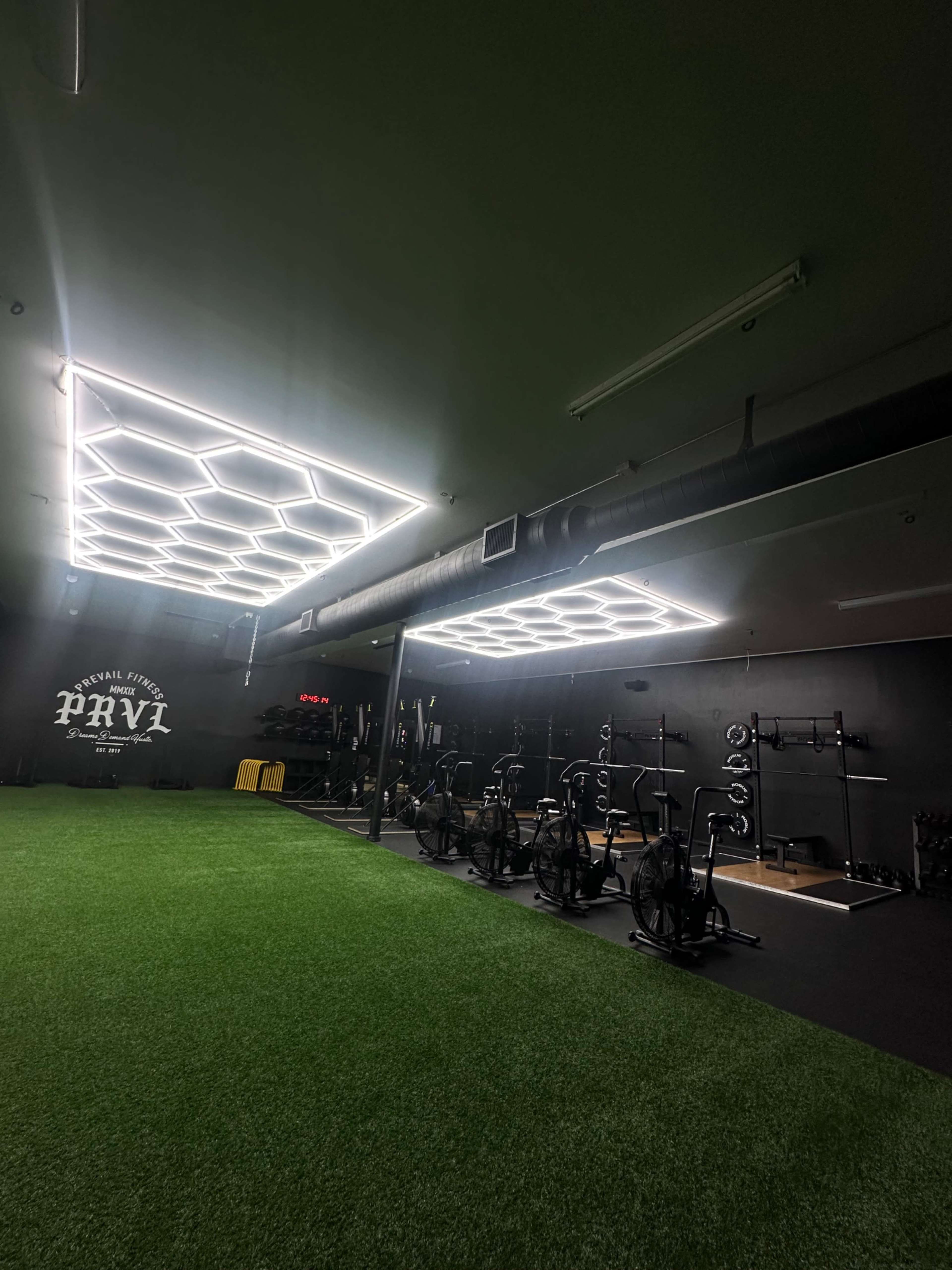 A modern gym space with hexagonal LED lighting, artificial grass flooring, and various fitness equipment arranged along the walls.