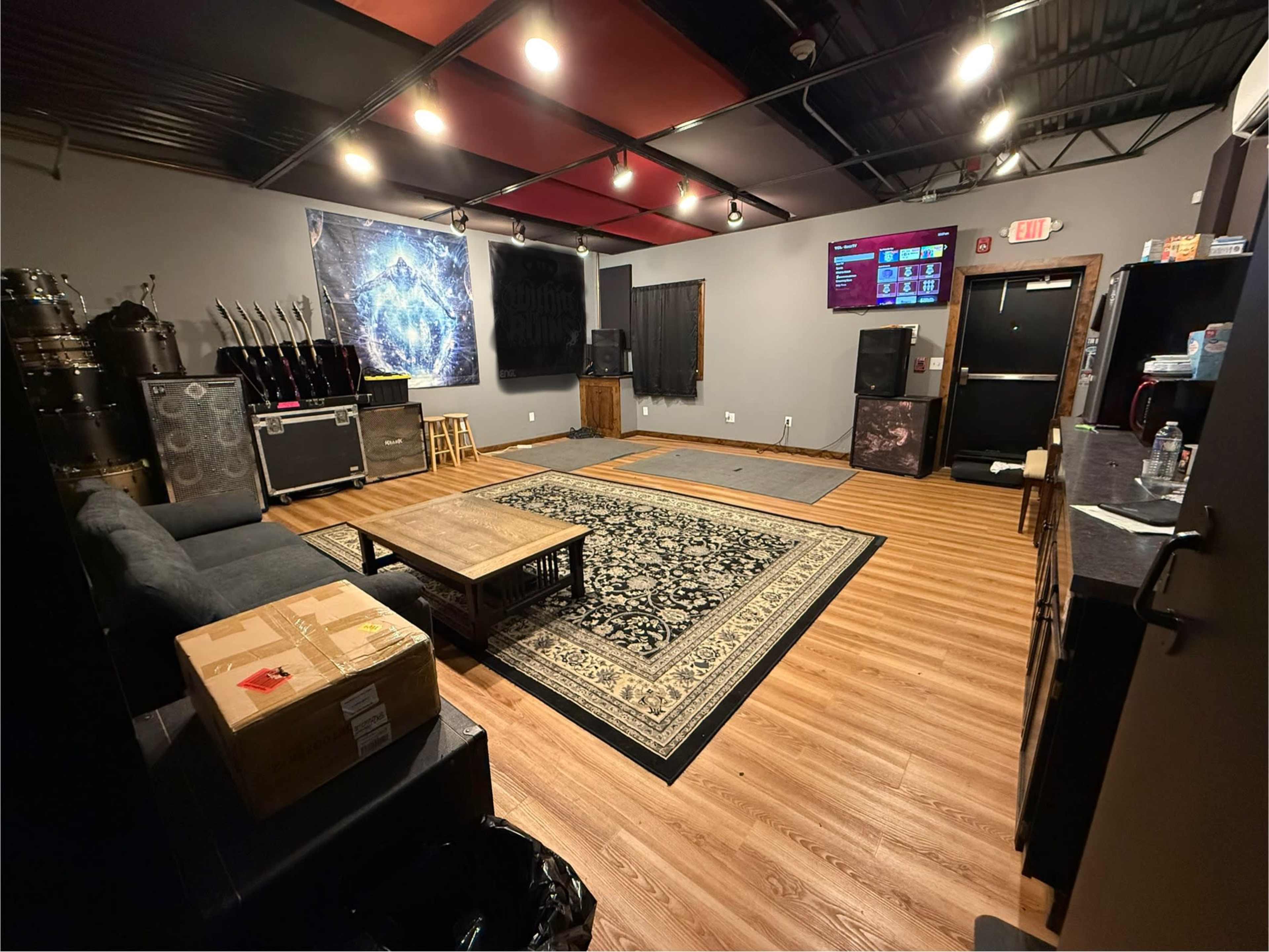 The image shows a music rehearsal space featuring a sofa, wooden coffee table, sound equipment, and a patterned rug on a wooden floor.