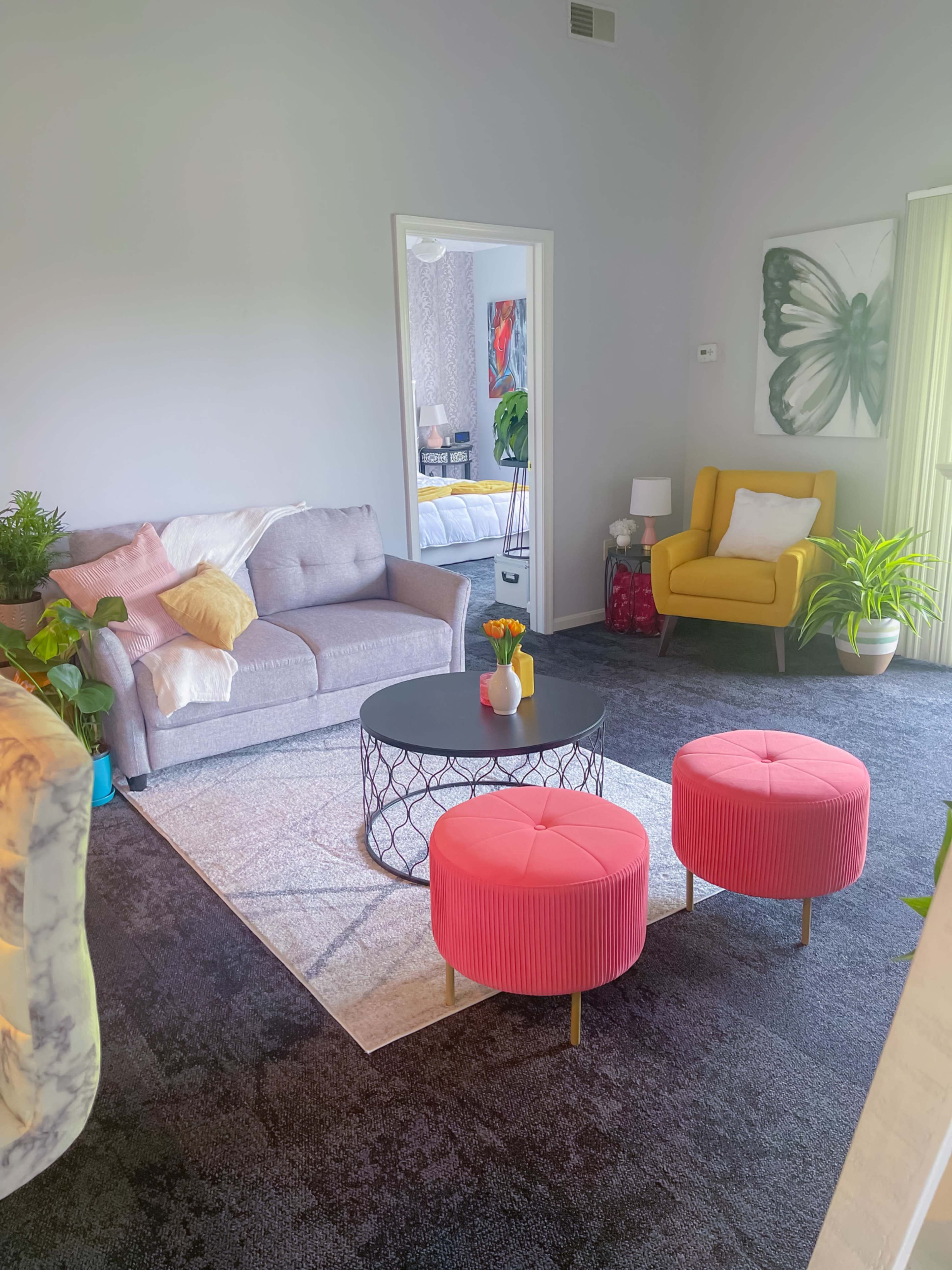 The living room features a gray sofa, a round coffee table, two pink ottomans, and various plants, creating a cozy atmosphere.