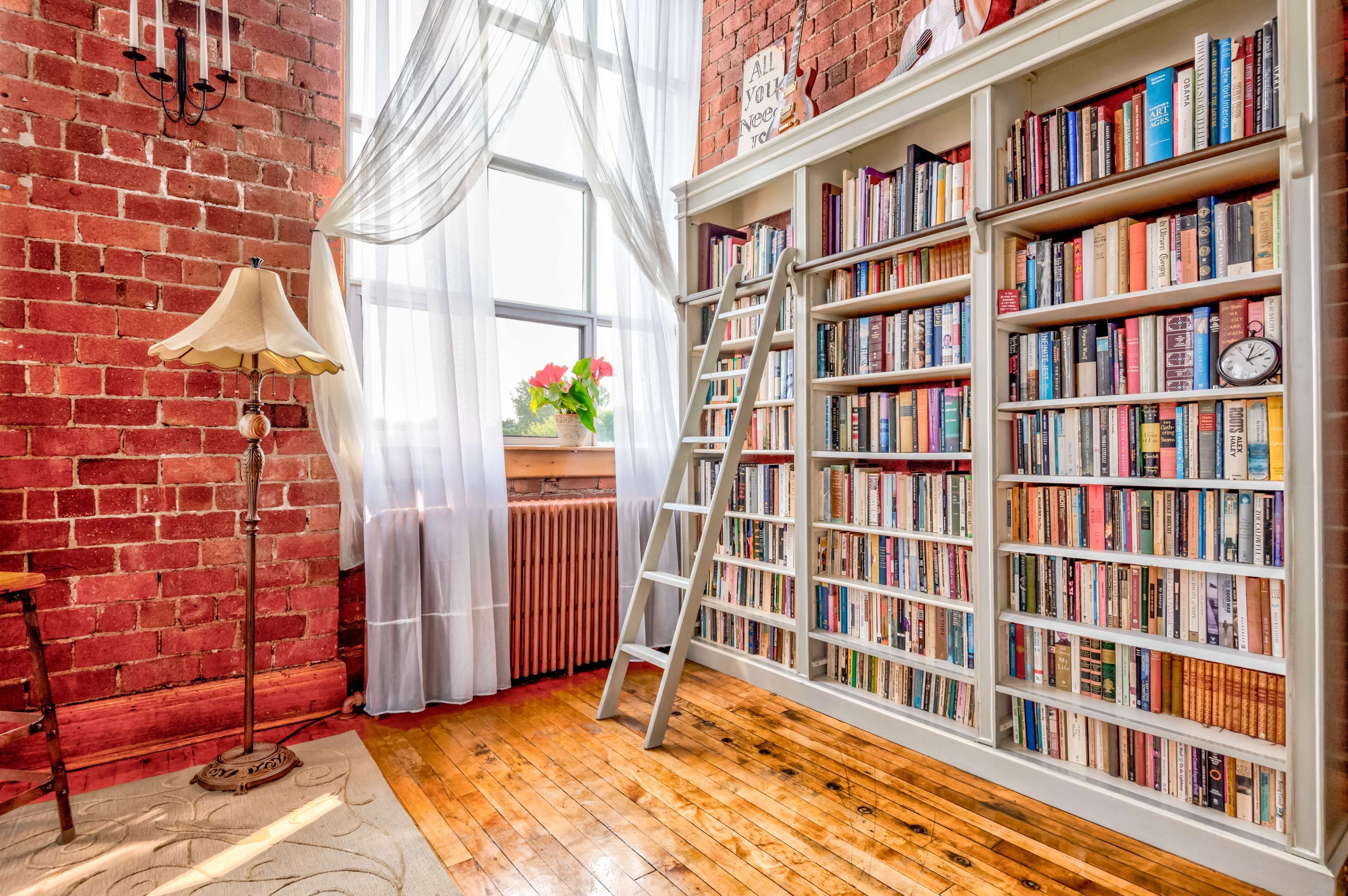 A cozy reading nook features a tall bookshelf filled with books, a metal ladder, a floor lamp, and large windows draped with sheer curtains.