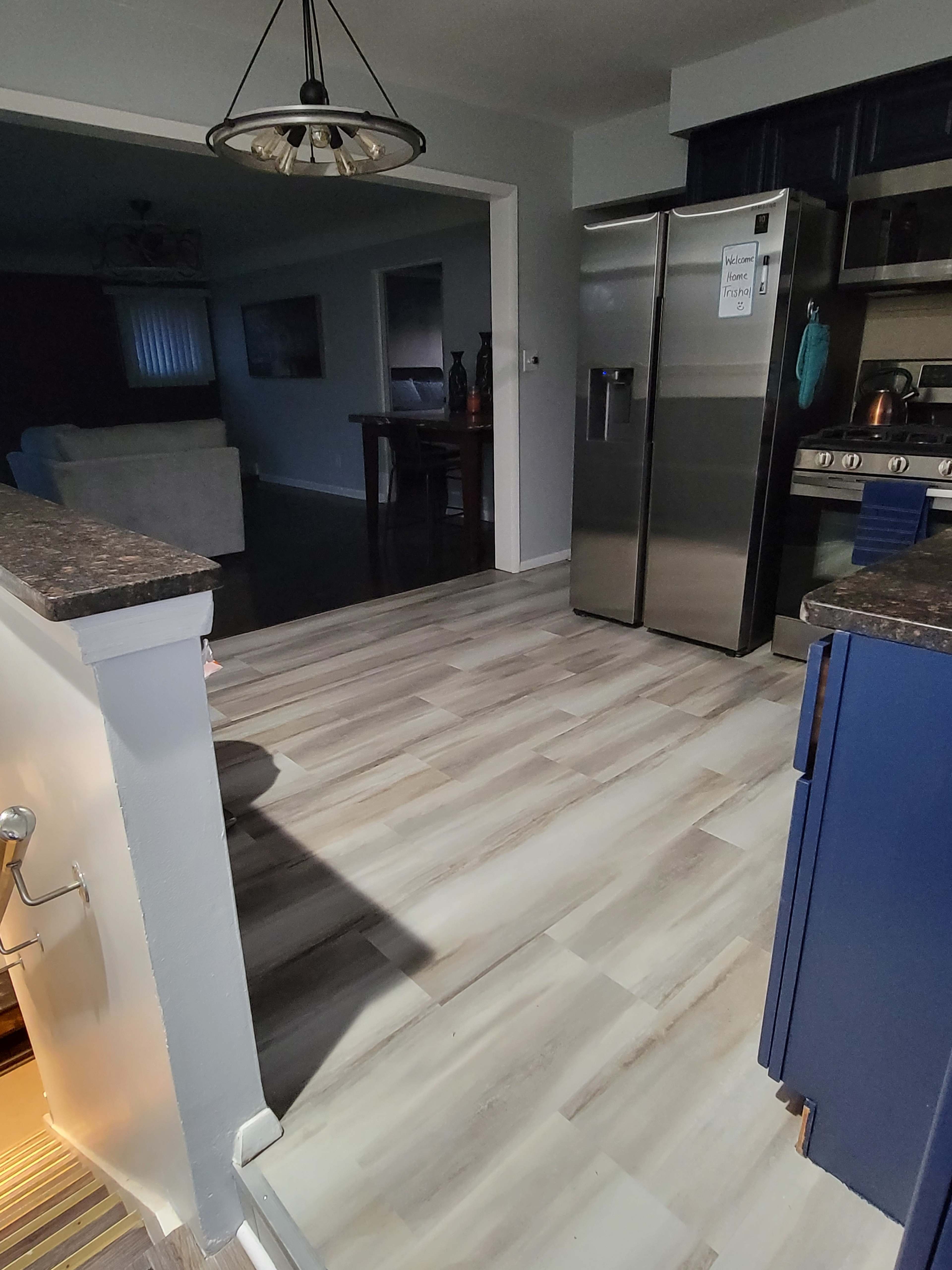 The image shows a kitchen area with stainless steel appliances, light-colored wooden flooring, and an adjoining living space visible in the background.