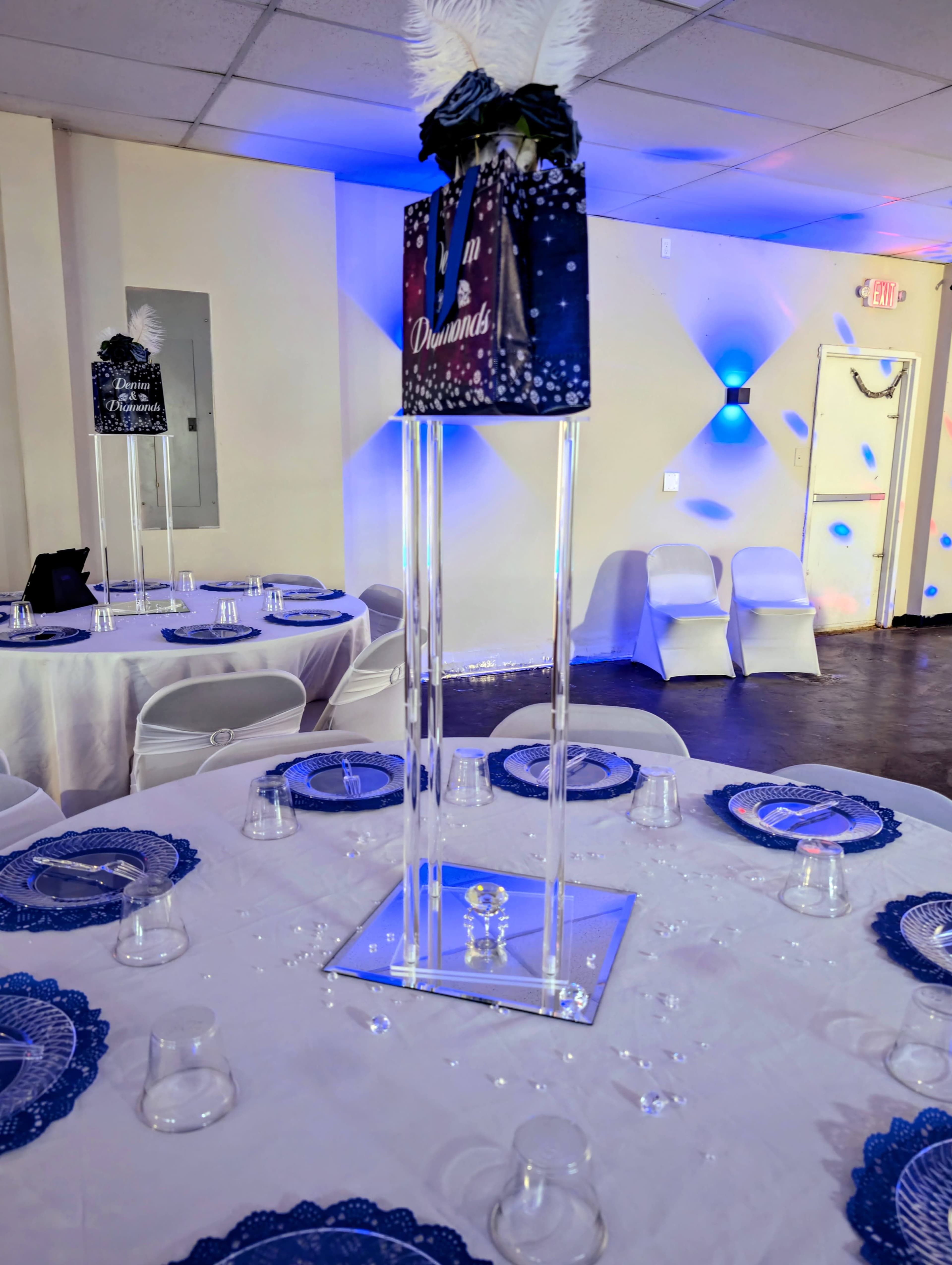 The image shows a banquet hall set up for an event, featuring round tables with white tablecloths and blue accents, clear glassware, and tall centerpieces on each table.