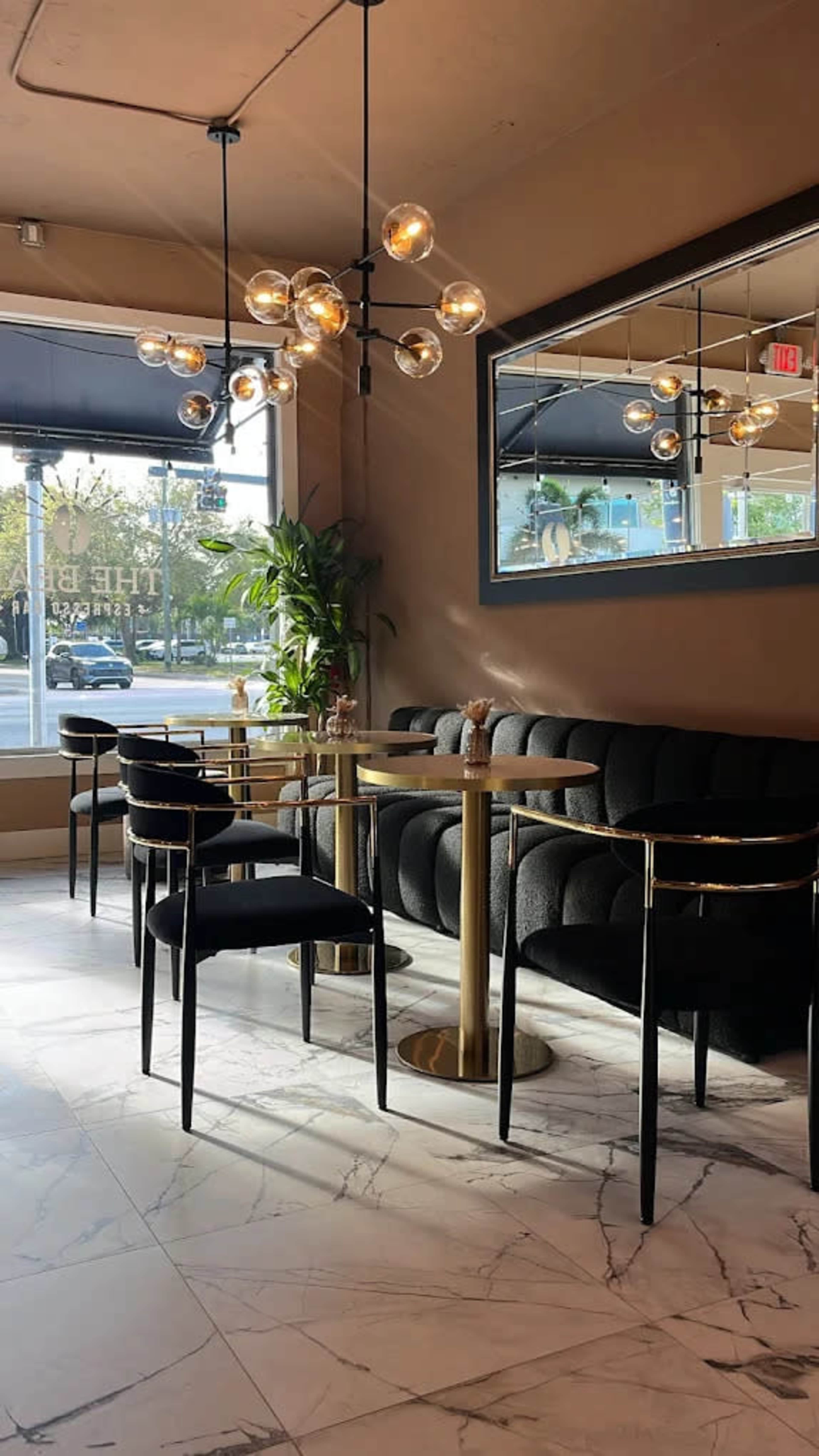 The image shows a modern cafe interior featuring a few black upholstered chairs and tables with gold accents, illuminated by a stylish chandelier and natural light coming through large windows.