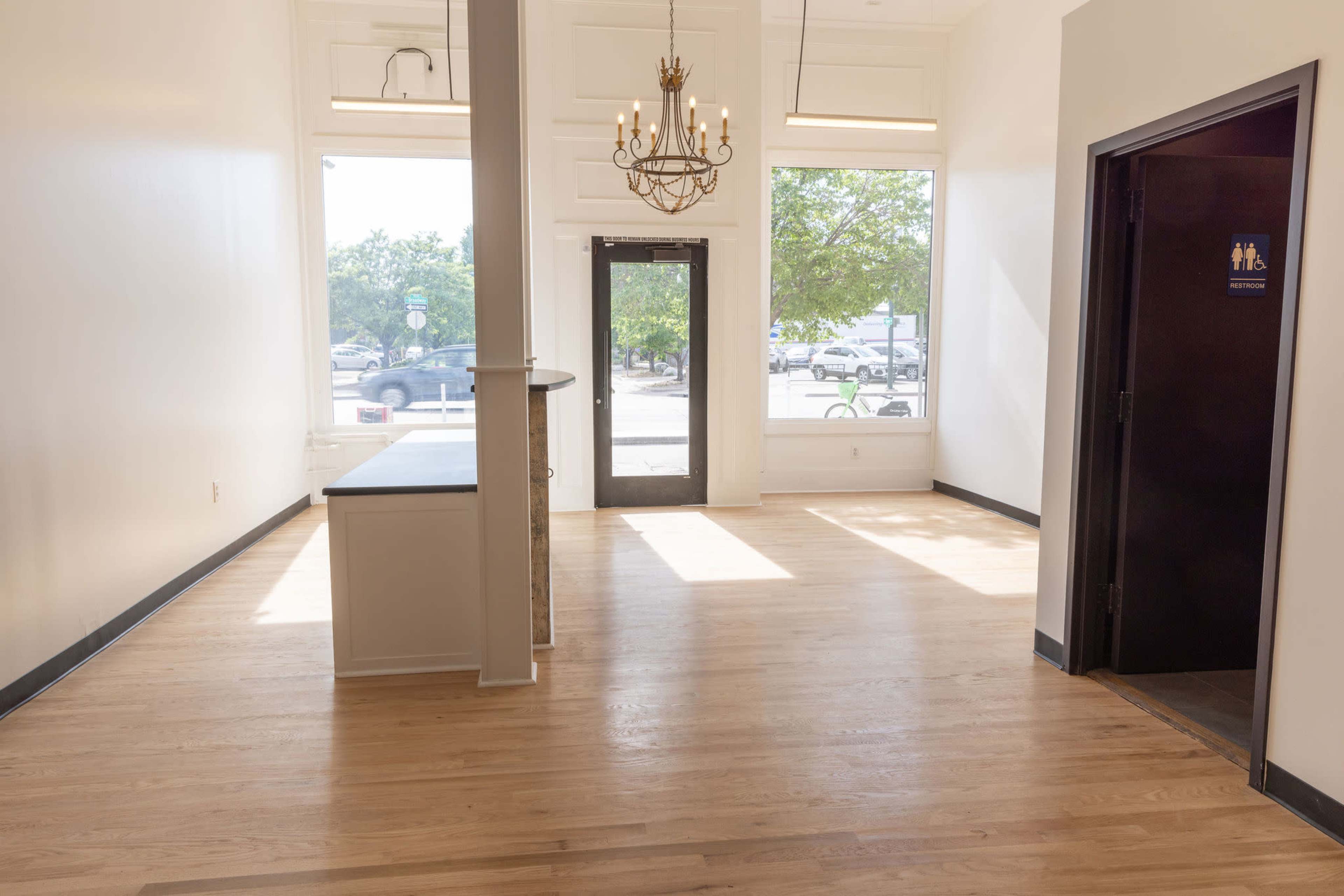 A vacant interior space featuring wooden flooring, large windows, a chandelier hanging from the ceiling, and a door labeled for restrooms.