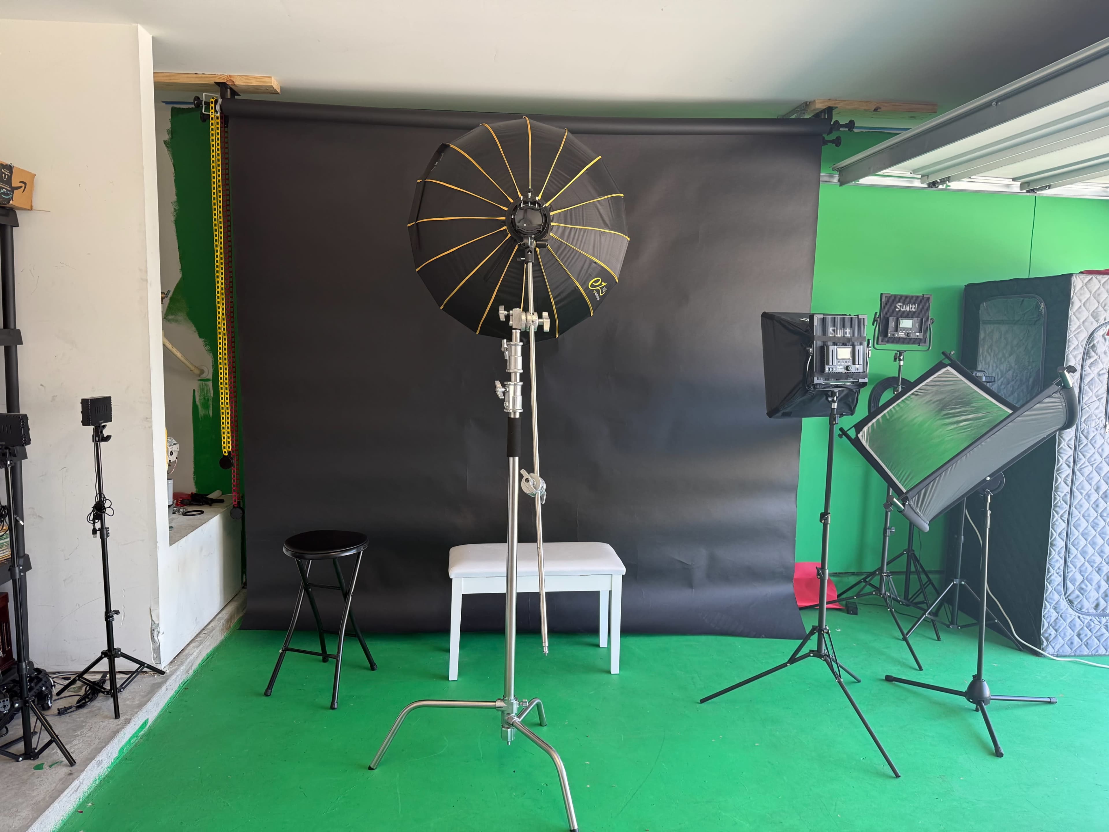 The image shows a photo studio setup with a black backdrop, various lighting equipment, a stool, and green flooring.
