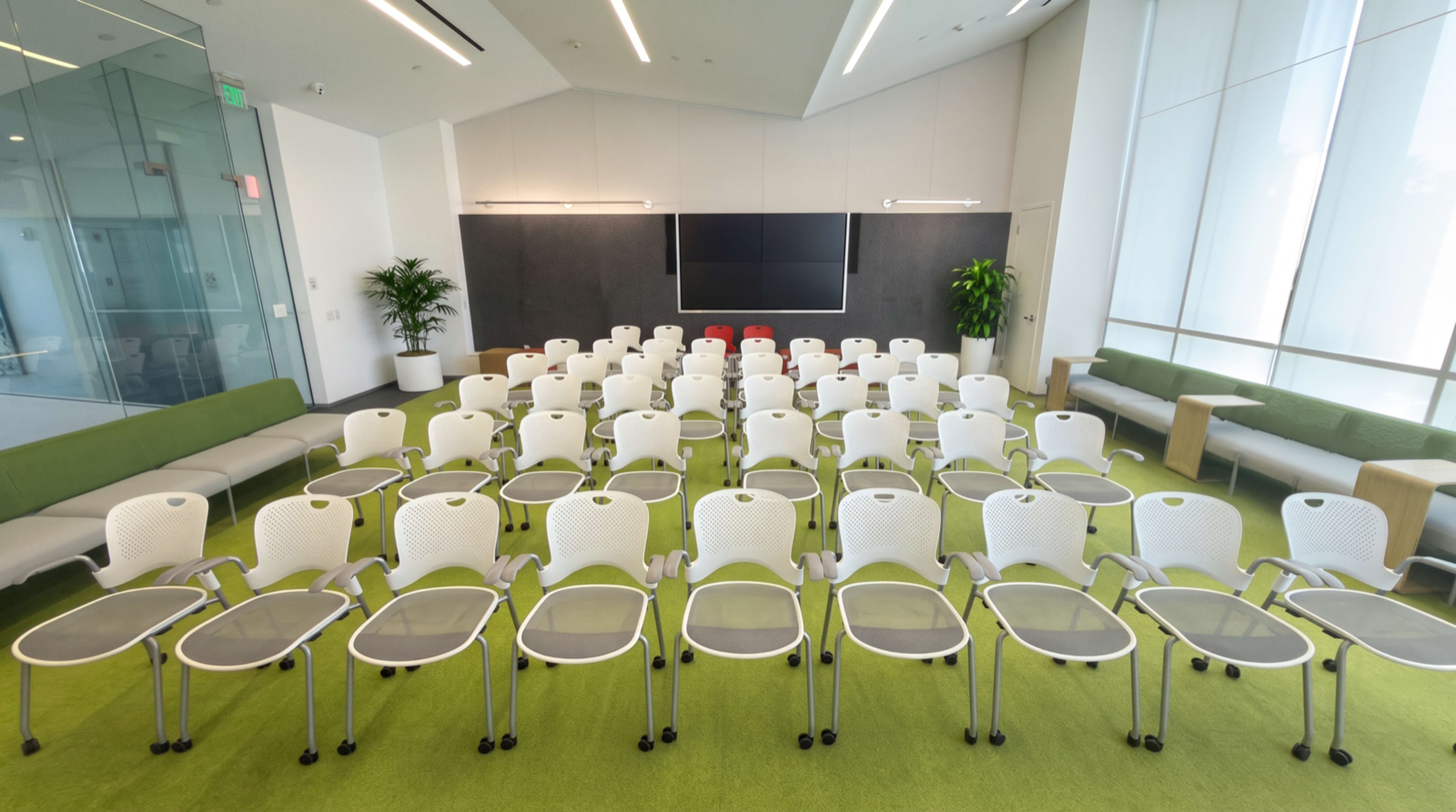 The image shows a modern conference room set up with rows of white chairs facing a large screen, surrounded by green carpeting and a few plants.