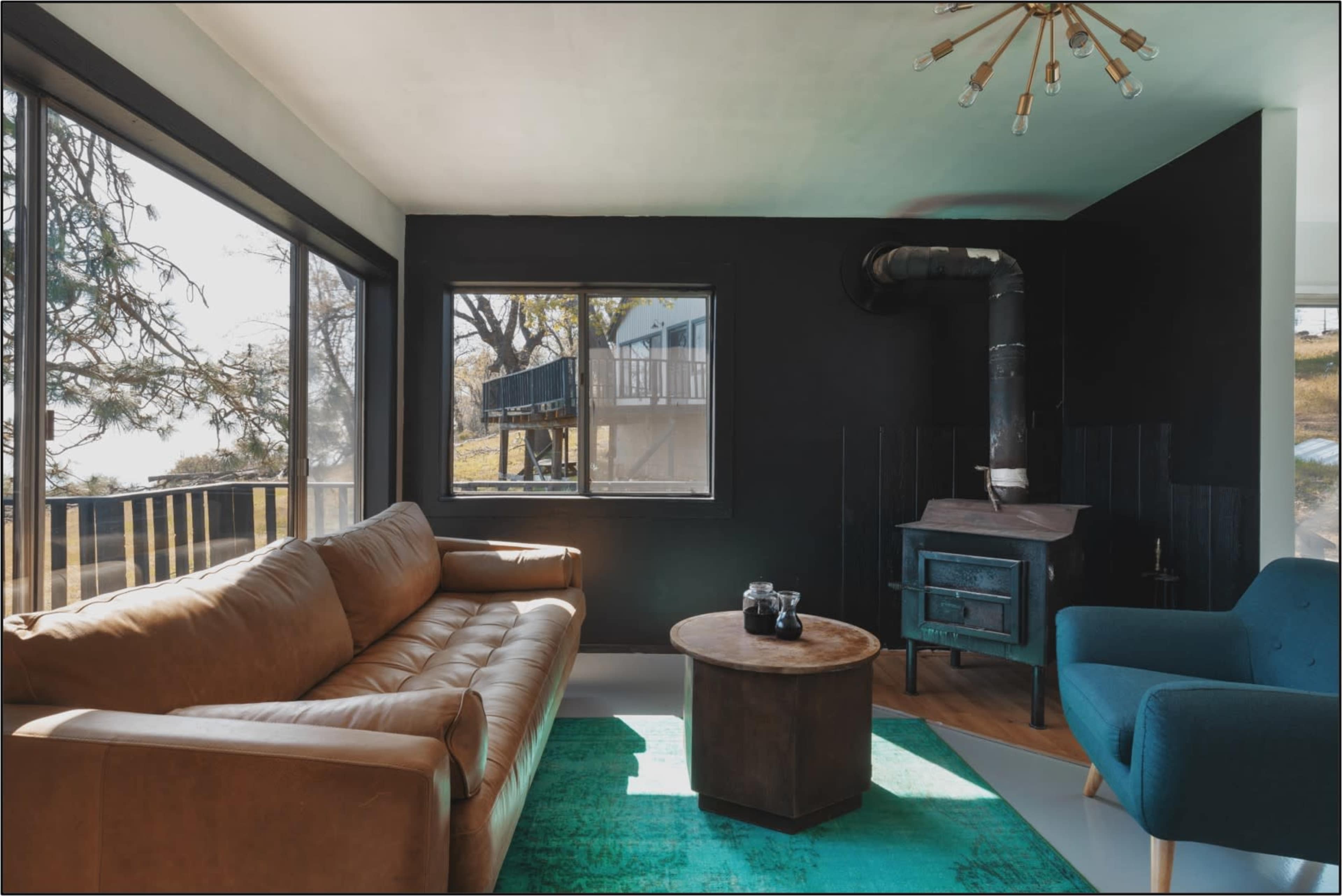The room features a brown leather sofa, a blue chair, a wooden coffee table, and a black-stove against a black wall, with large windows allowing natural light to enter.