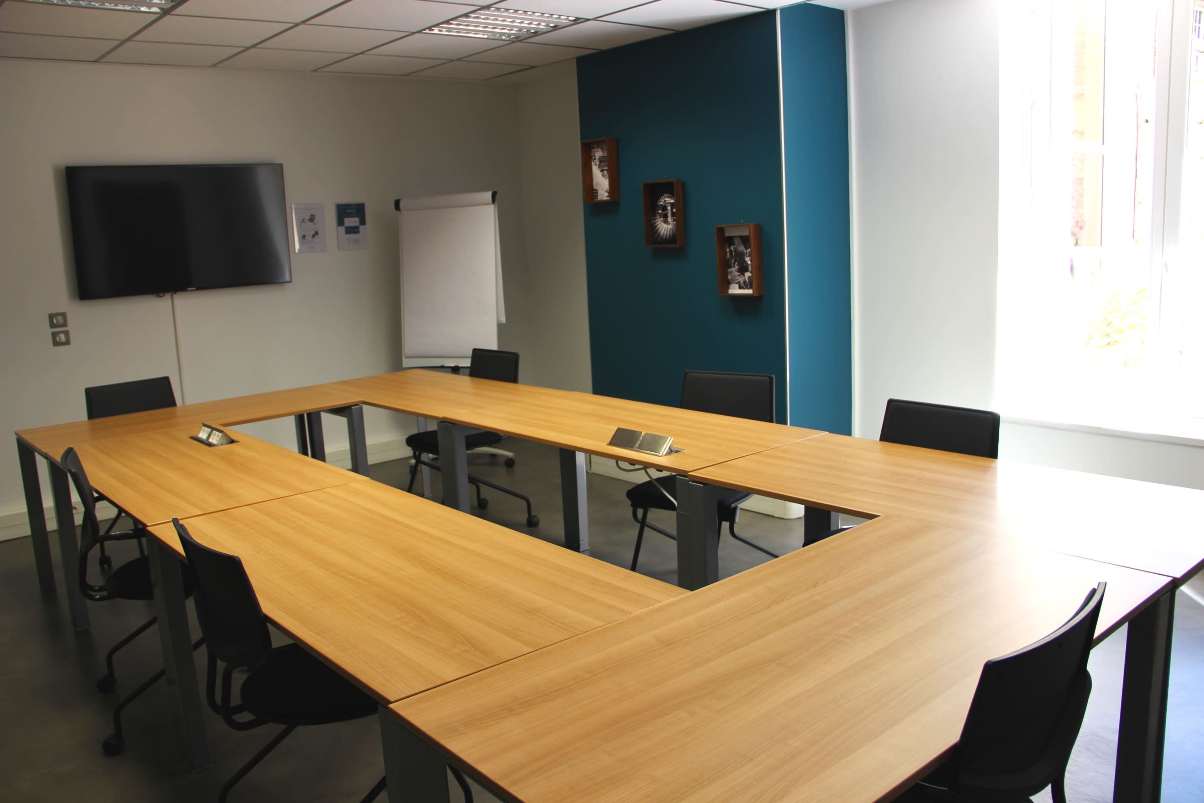 A conference room with a wooden table arranged in a U-shape, surrounded by black chairs, and equipped with a TV screen and a flip chart.