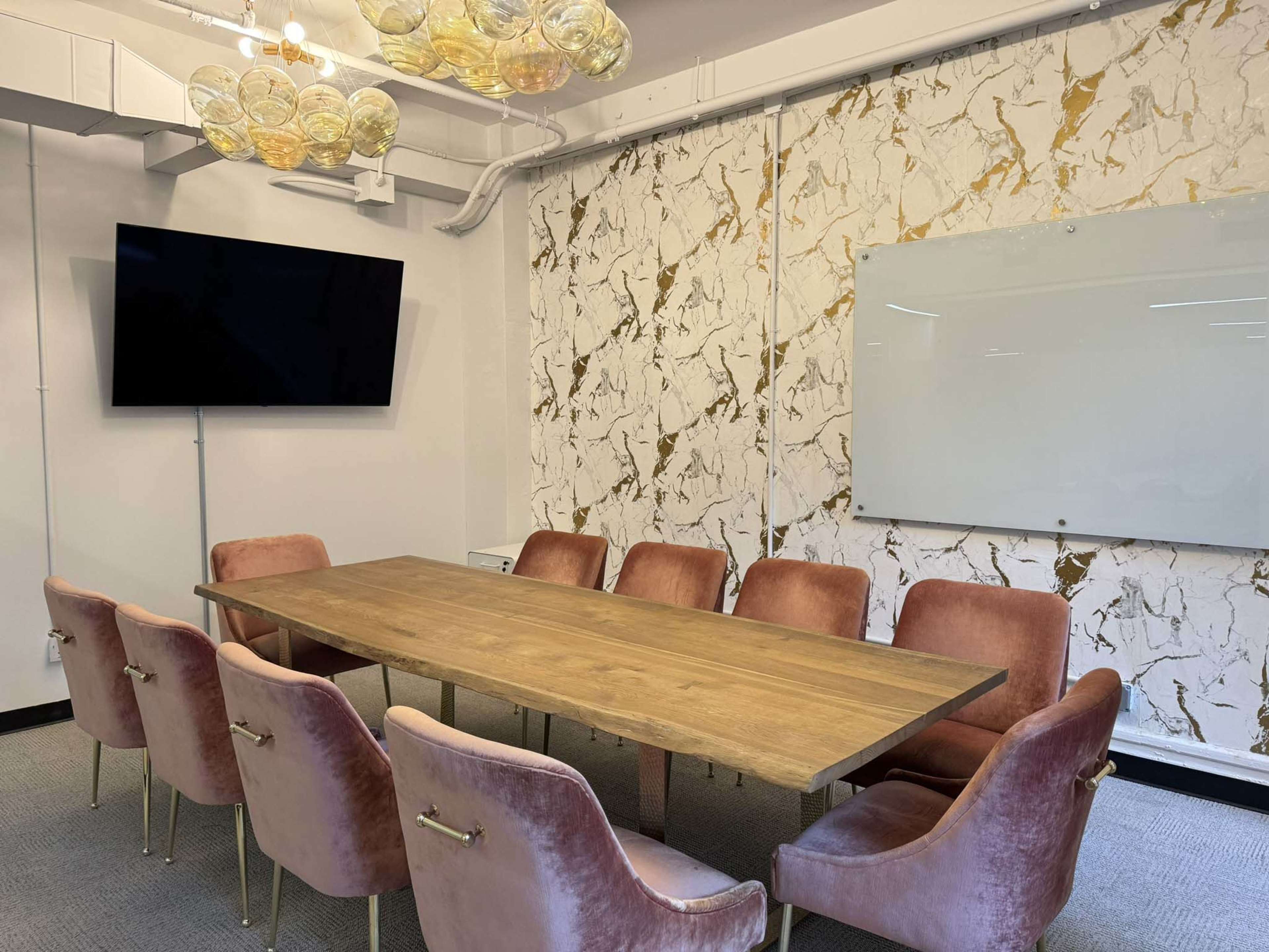 The image shows a modern conference room featuring a long wooden table surrounded by pink upholstered chairs, a large television mounted on the wall, and a whiteboard beside a decorative marble-patterned wall.