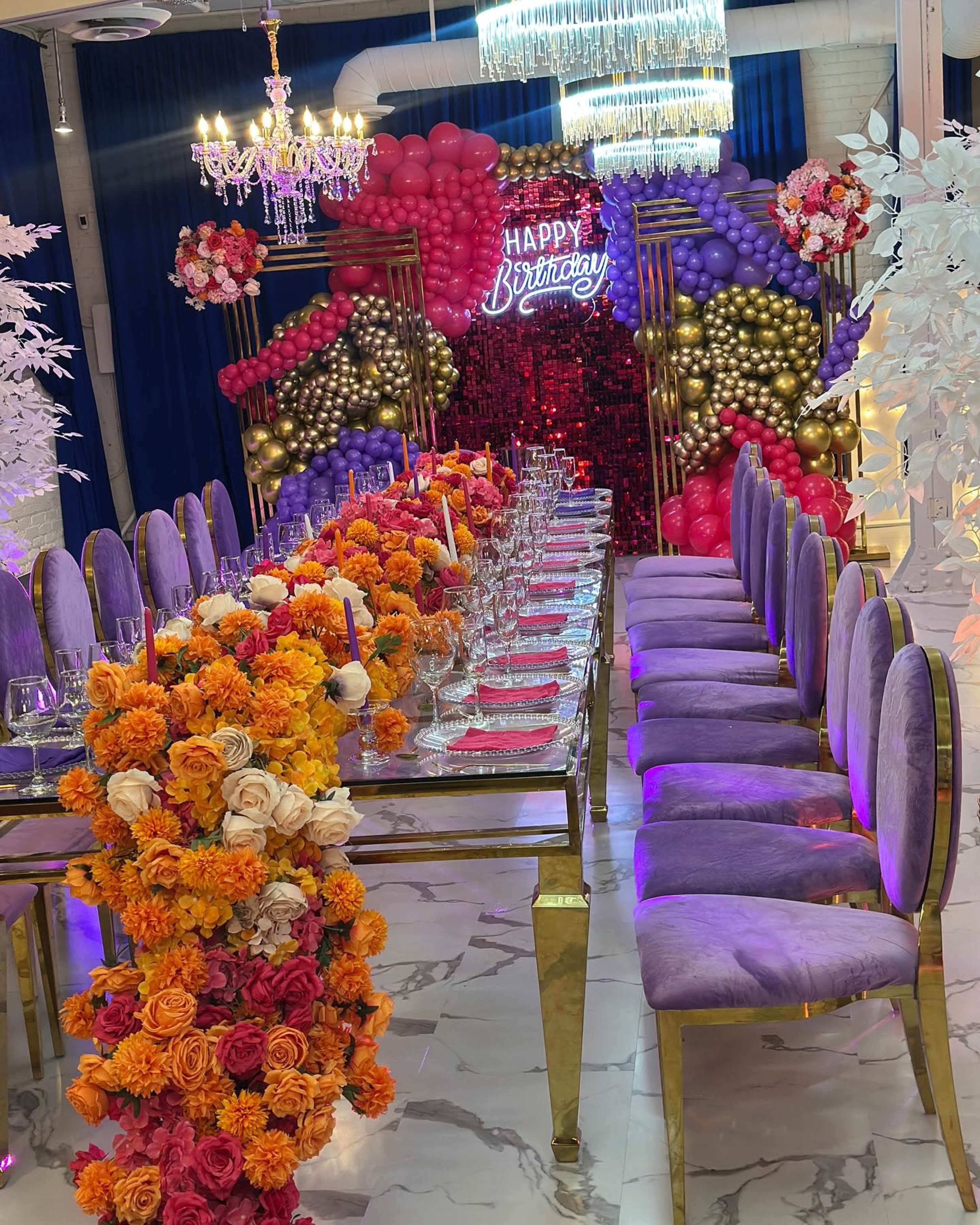 A long table adorned with vibrant flower arrangements and surrounded by plush purple chairs is set against a festive backdrop of balloons and a "Happy Birthday" sign.