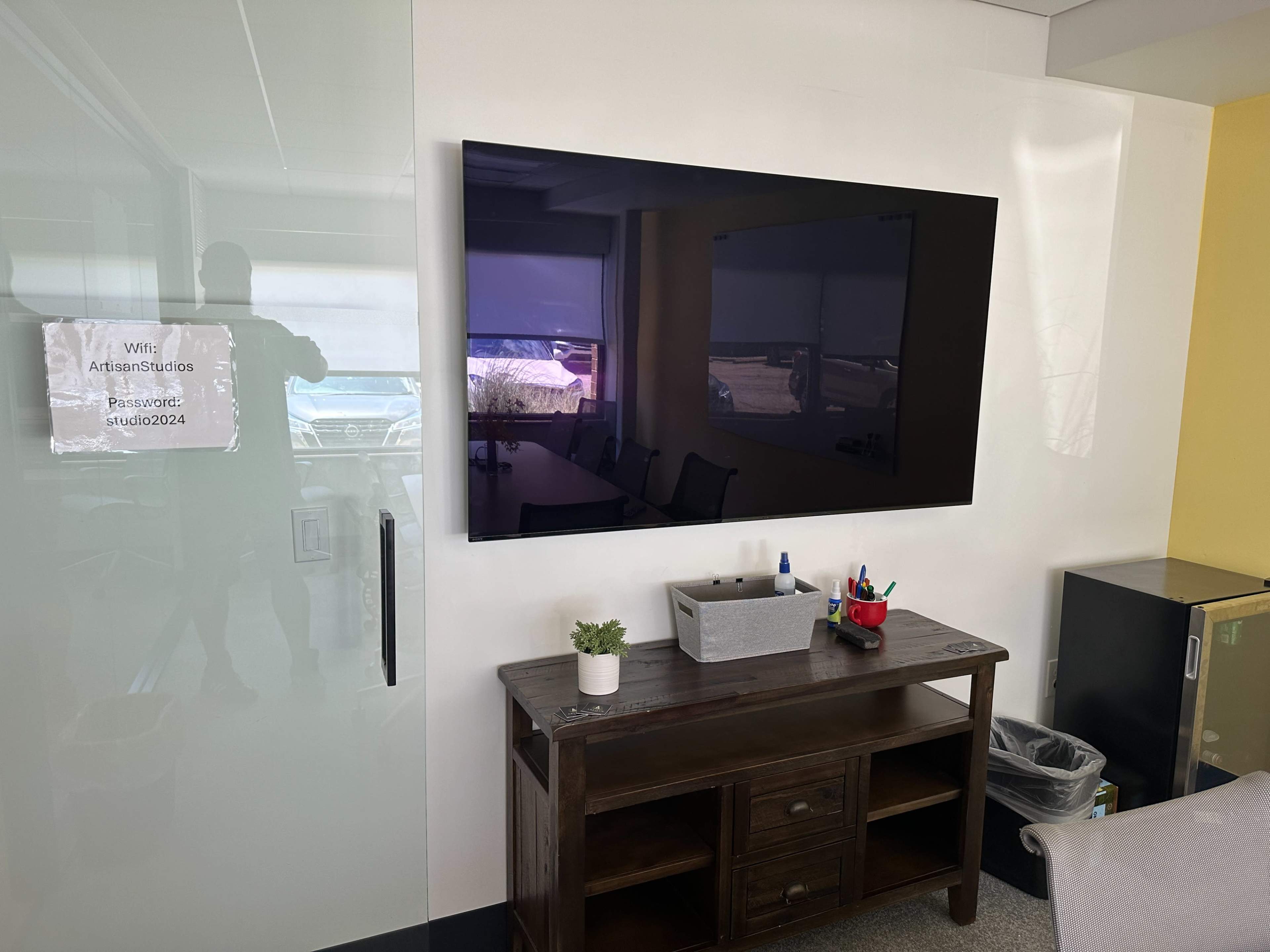 A modern office space featuring a large flat-screen TV mounted on a white wall, alongside a dark wooden console table with a small plant and various office supplies.