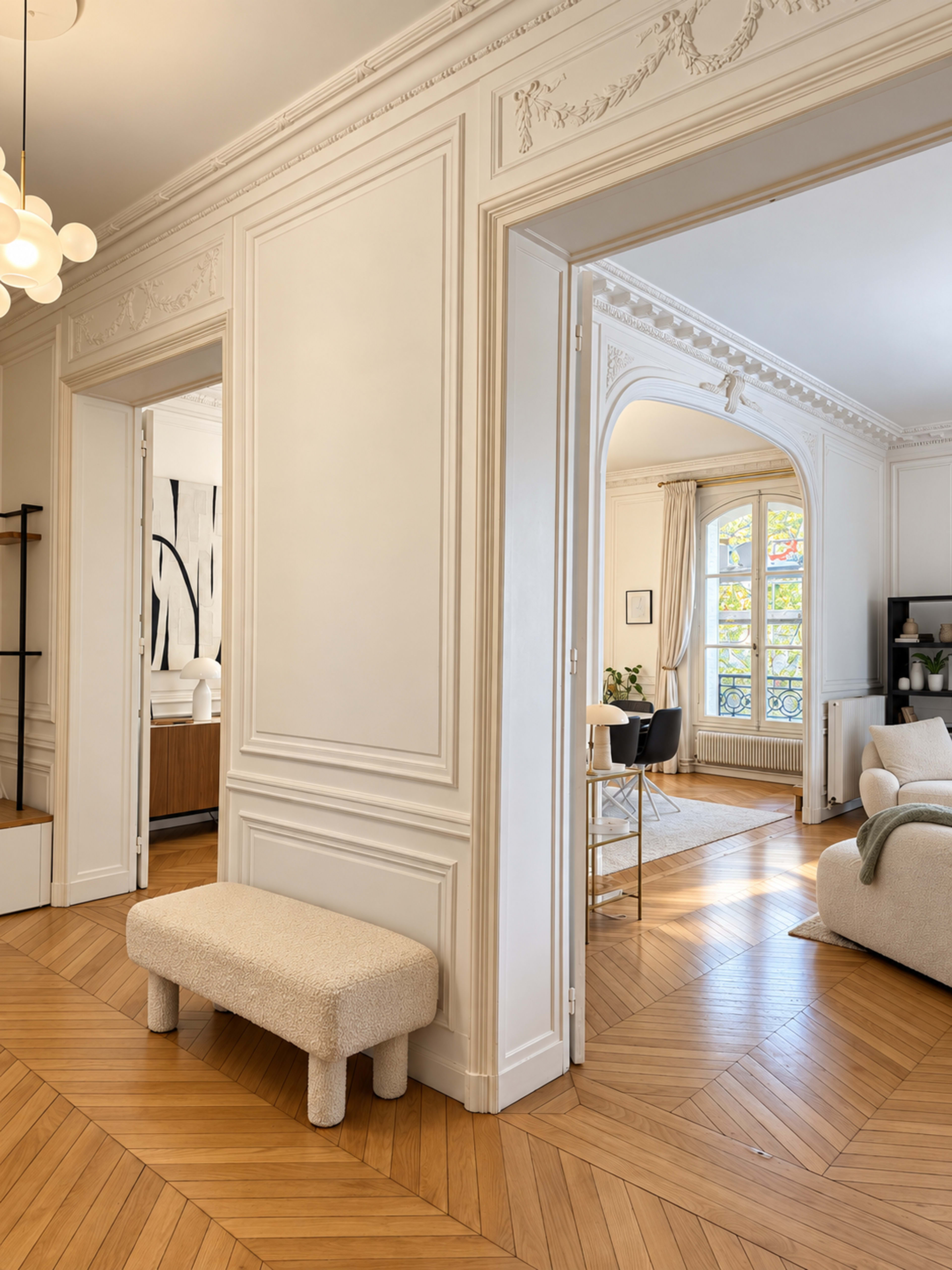 The image shows a bright interior space featuring intricately detailed white walls, a cozy seating area, and a view into an adjoining room with wooden flooring.