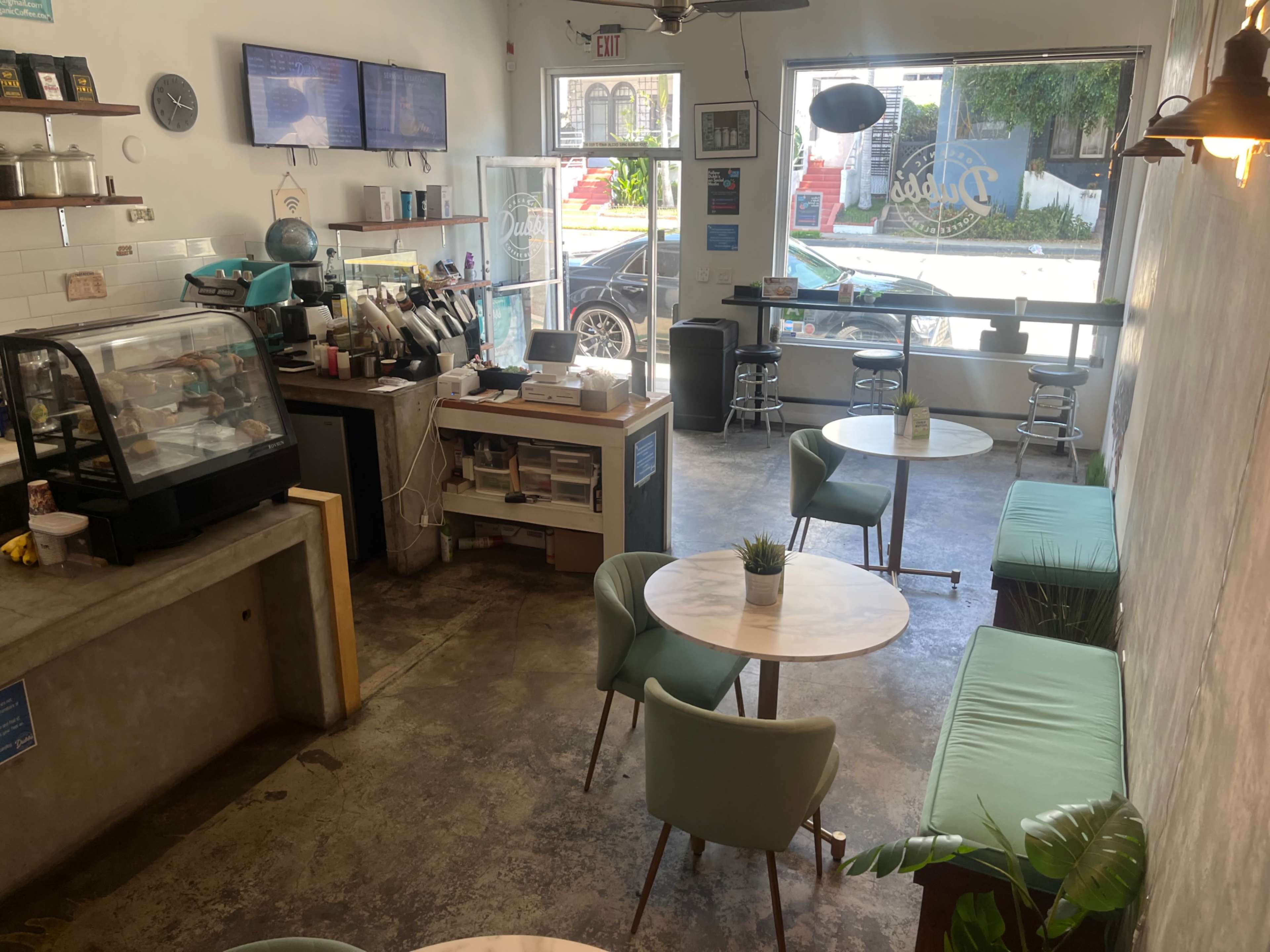 The image shows a cozy café interior with green seating, two marble-topped tables, a display case for baked goods, and a coffee preparation area near a large window.