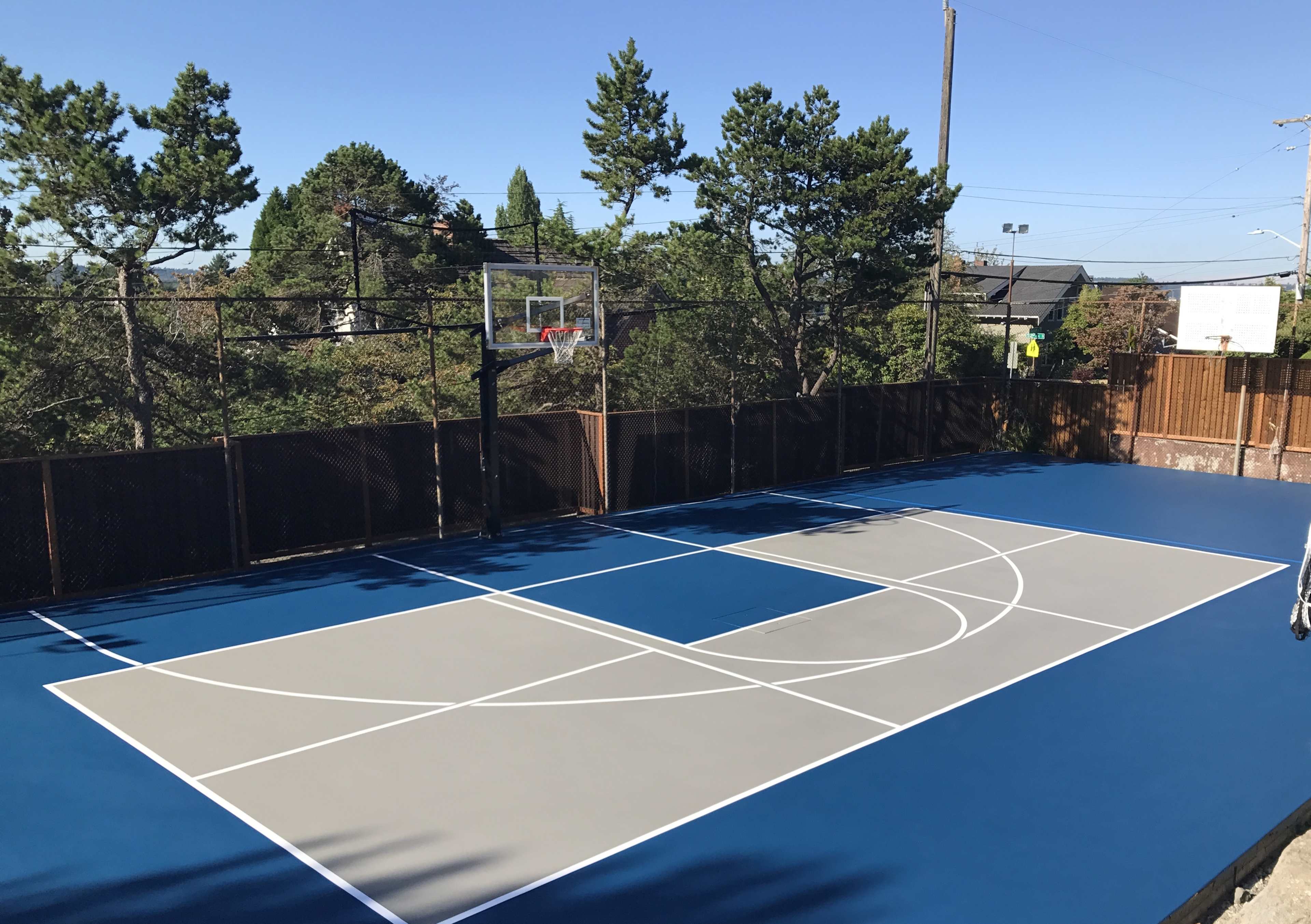 A newly constructed basketball court with blue and gray paint, surrounded by black fencing and trees in the background.