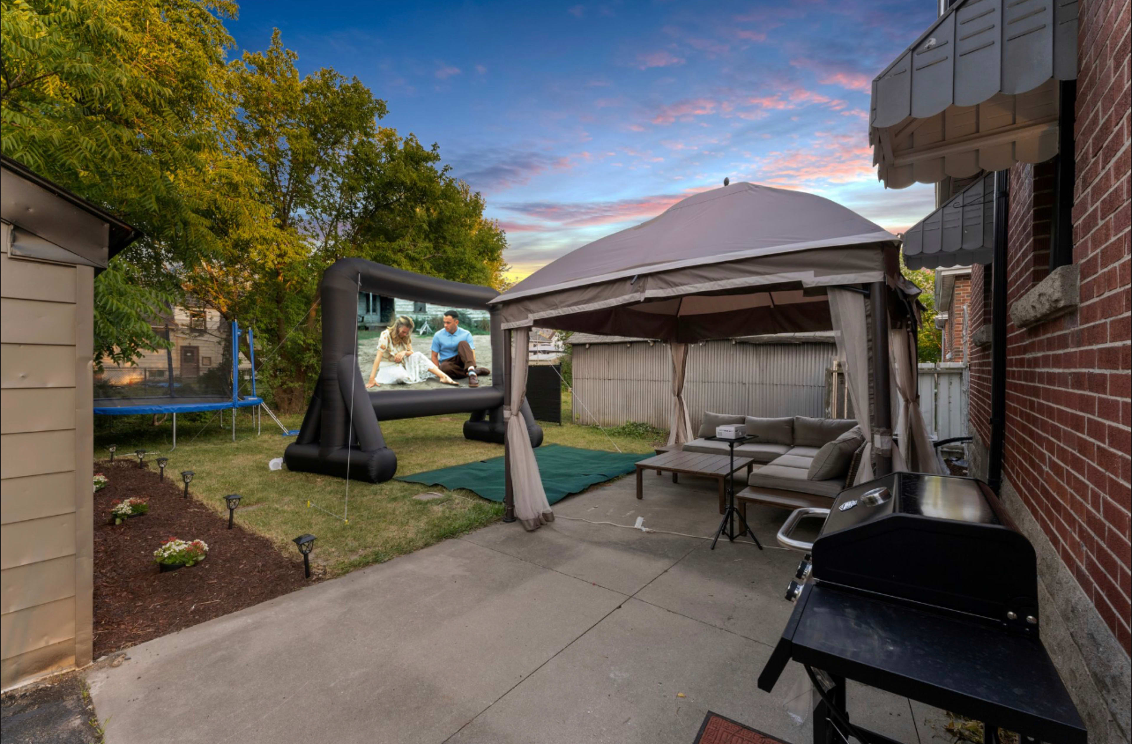 A backyard scene features a gazebo, outdoor seating, a grill, a trampoline, and a large inflatable screen displaying a video, set against a colorful sky.