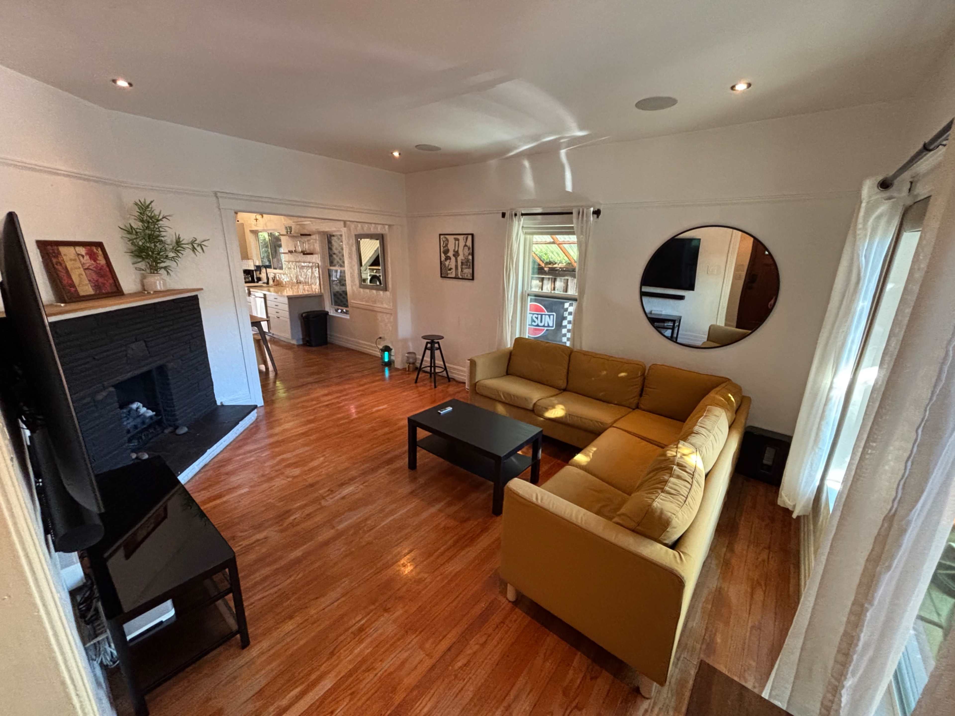 The image shows a spacious living room with a yellow sectional sofa, a dark coffee table, and a flat-screen TV against a wall with hardwood flooring.