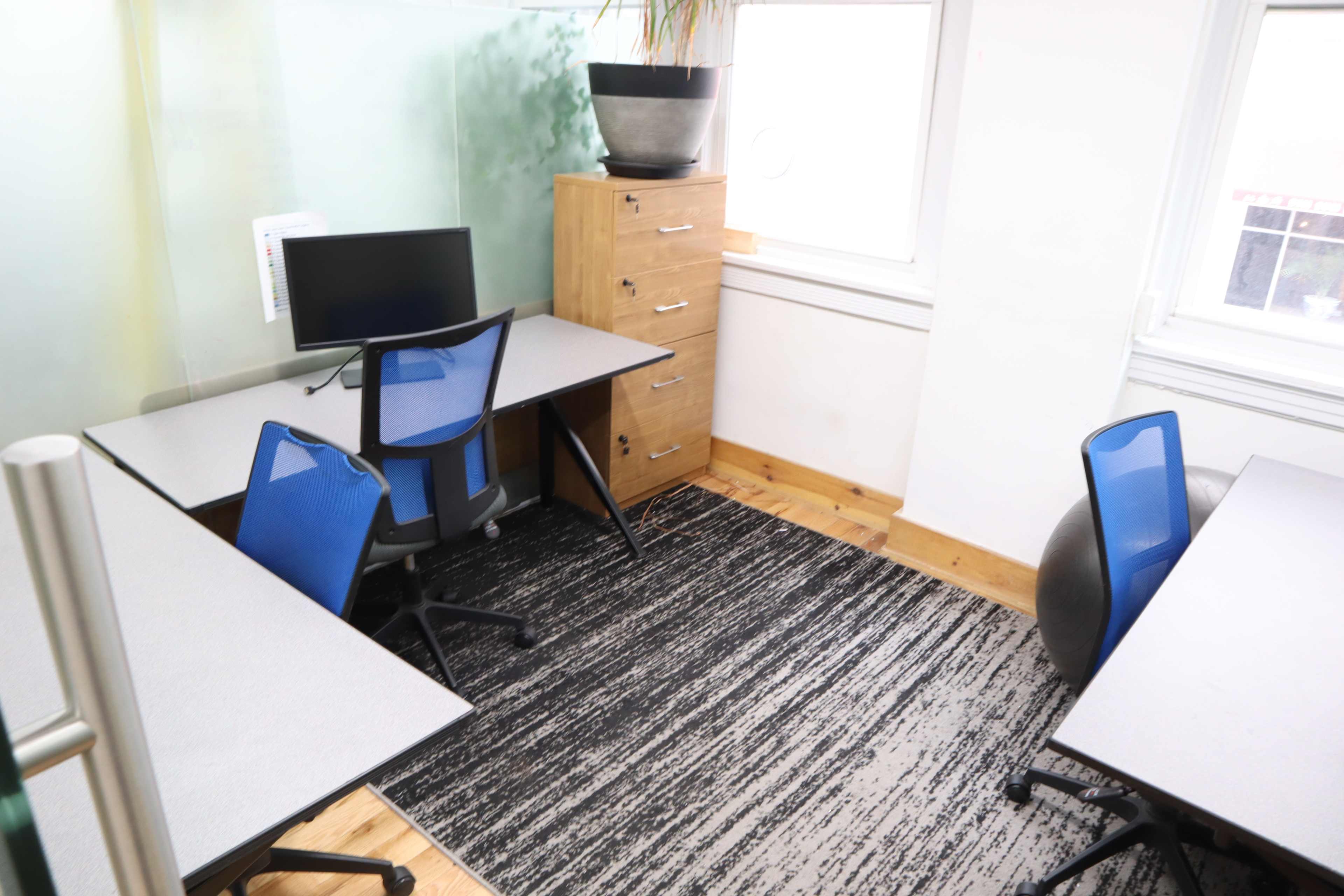 A small office space with two desks, each equipped with blue chairs, a computer monitor, and a filing cabinet against the wall.