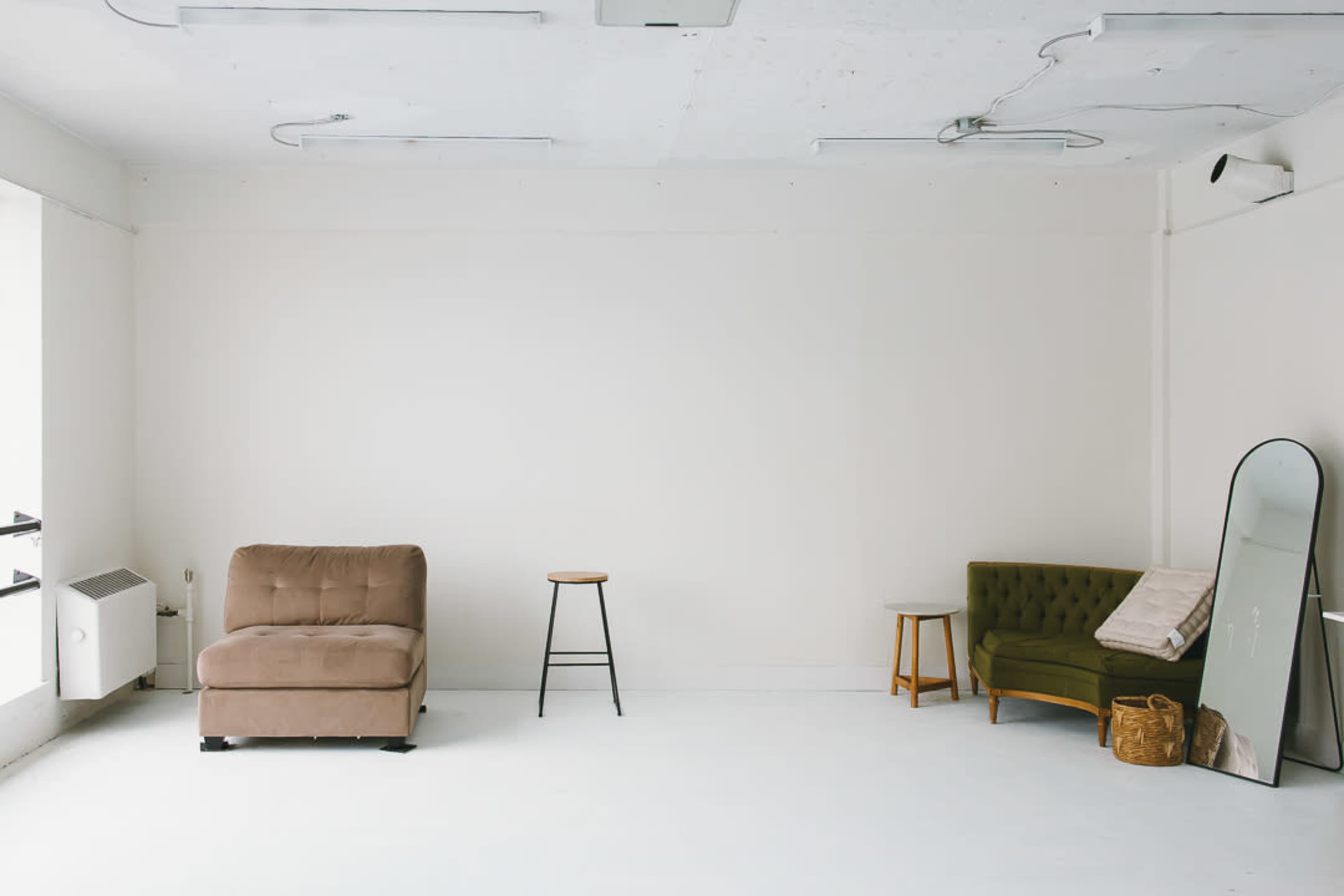 The image shows a minimalist interior space with a beige couch, a green sofa, a small round table, a wooden stool, and a large mirror against a white wall.