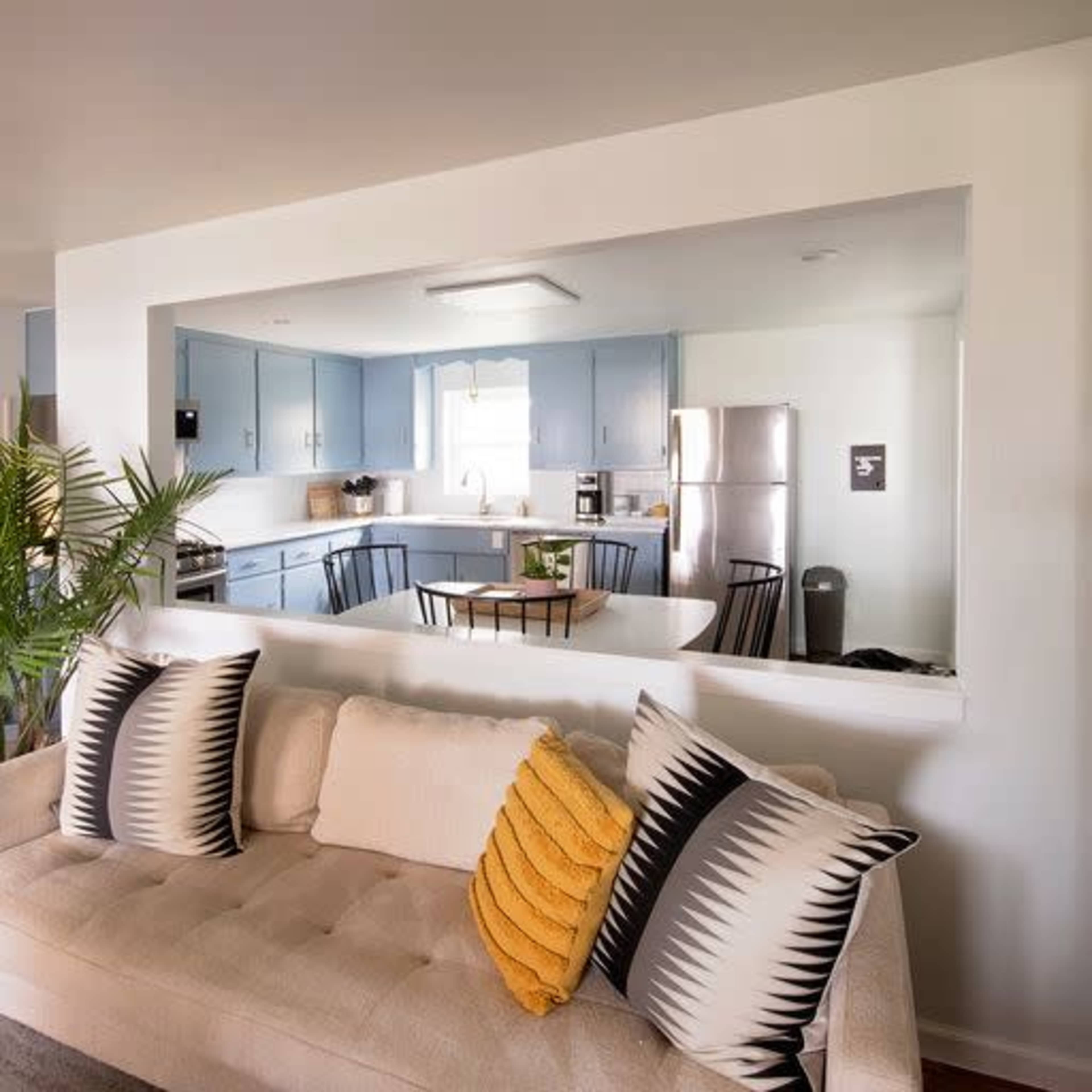 A living area featuring a light-colored sofa with patterned cushions, a dining table in the adjoining kitchen, and modern appliances in a bright, open layout.