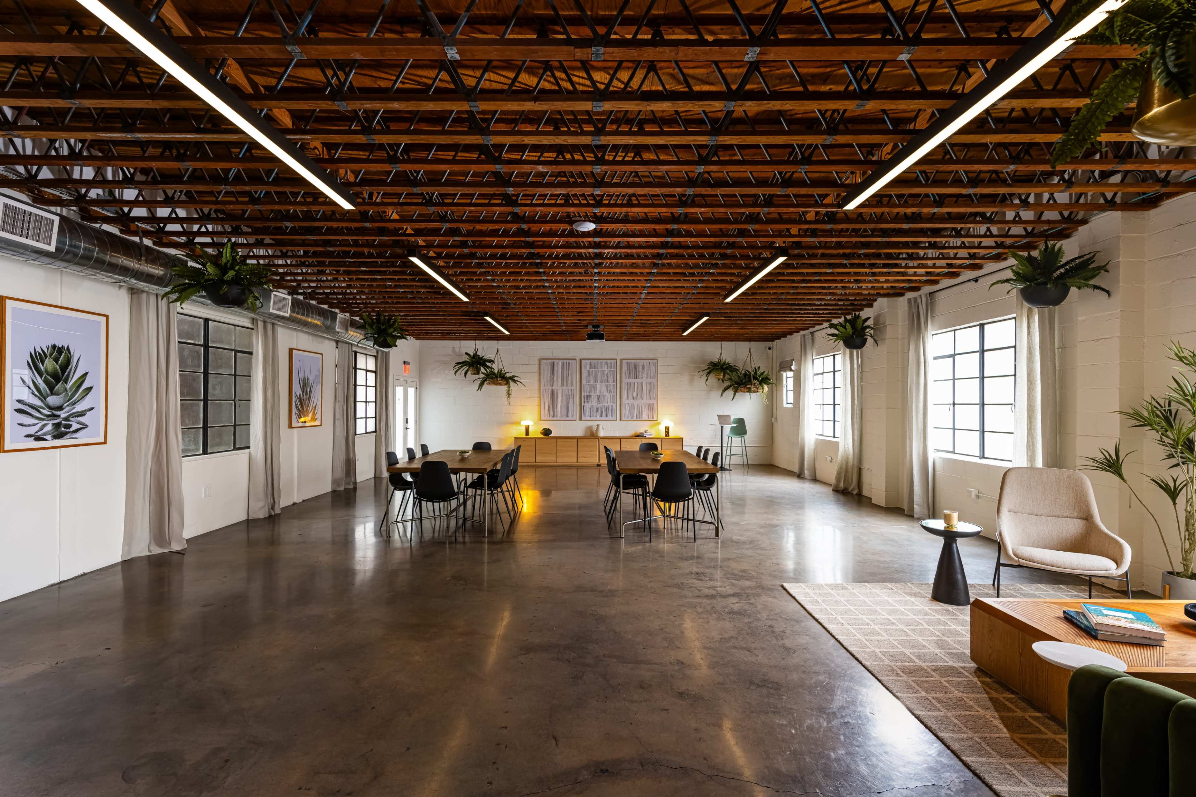 The image depicts a spacious, well-lit room featuring a meeting area with tables and chairs, wooden beams overhead, and decorative plants hanging from the walls.