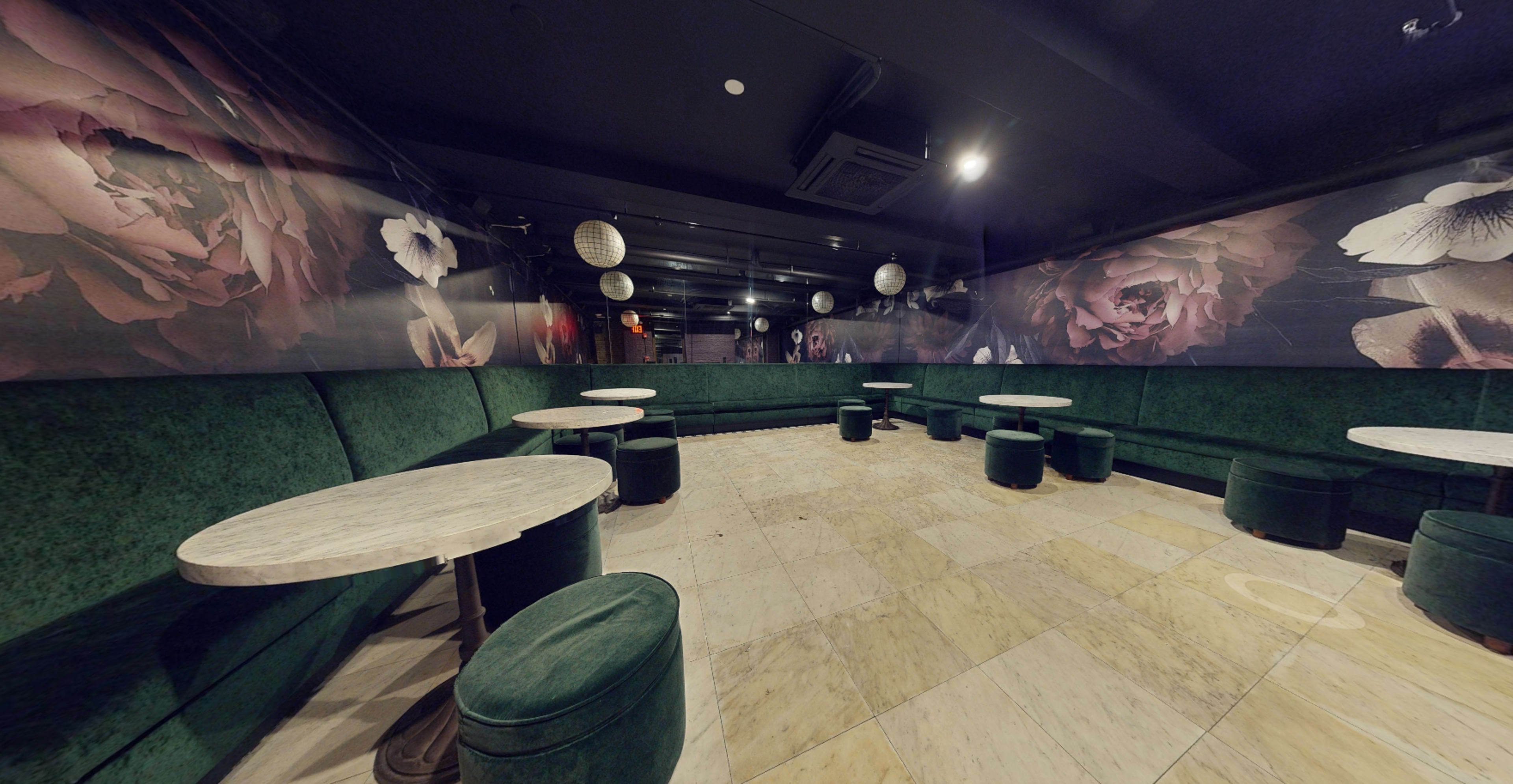 The image shows a dimly lit lounge area featuring green upholstered seating, round tables, and floral wall decor.