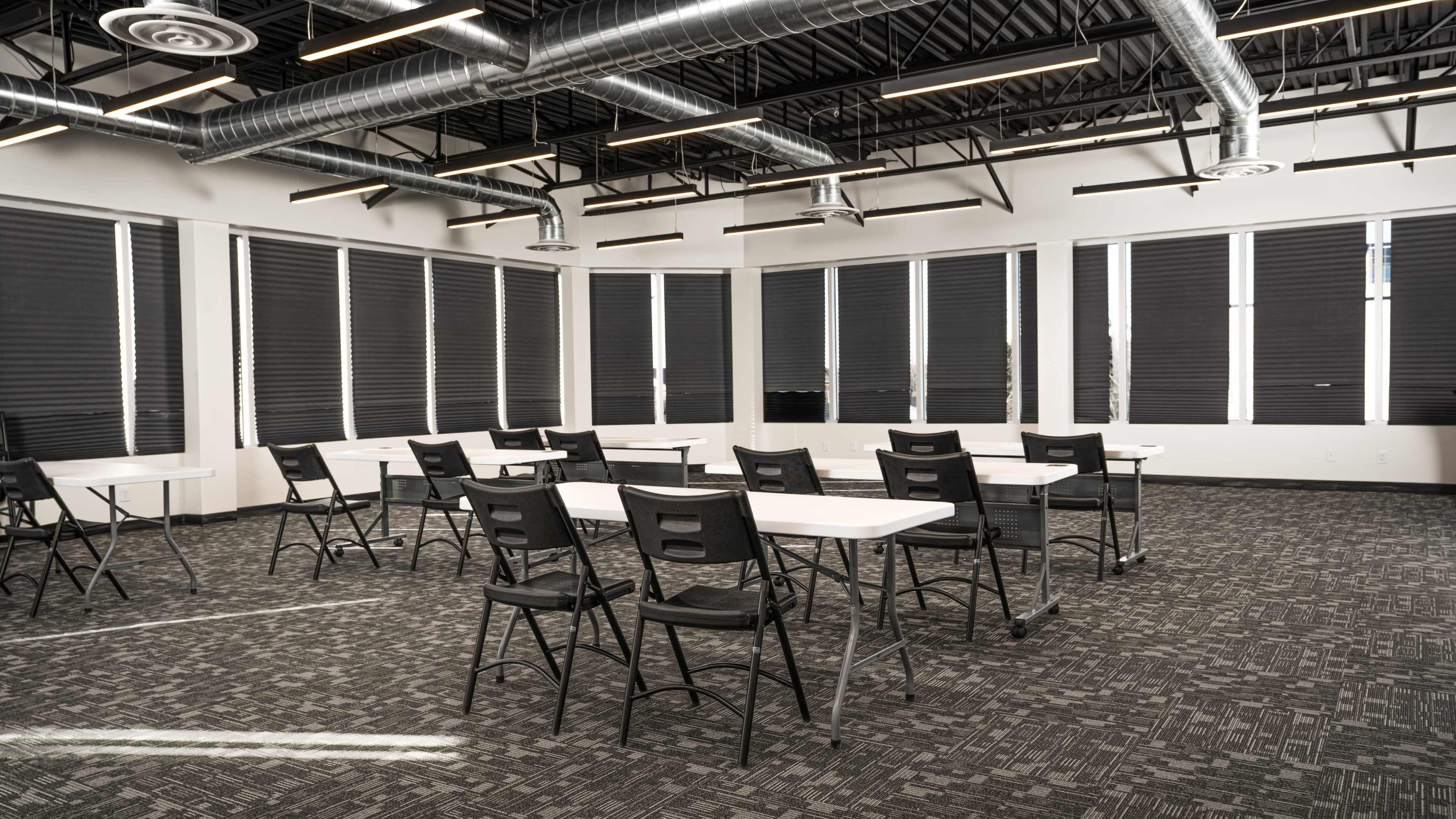 A classroom features several rows of white tables and black chairs arranged on a carpeted floor, with large windows covered by retractable blinds.