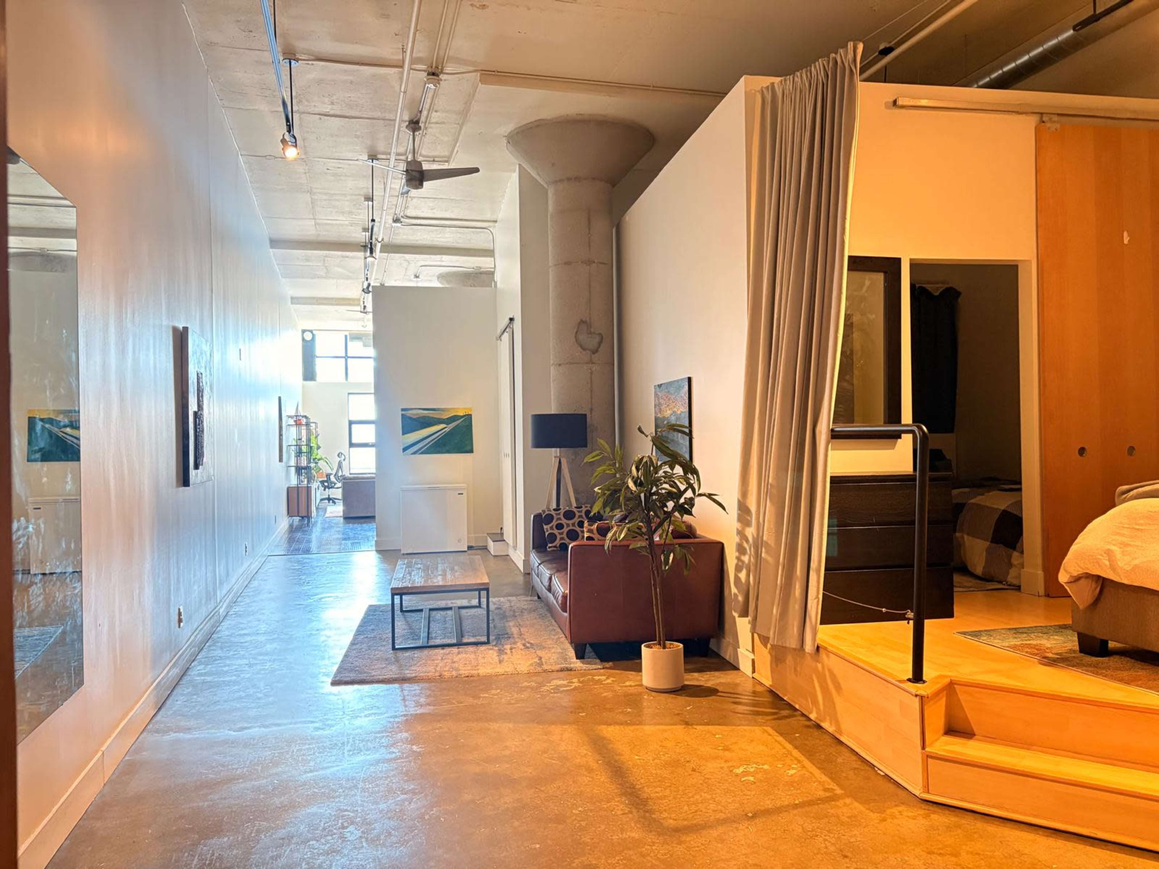 The image shows a spacious interior featuring a modern living area with a brown sofa, a small coffee table, a partitioned sleeping area, and large windows allowing natural light to enter.